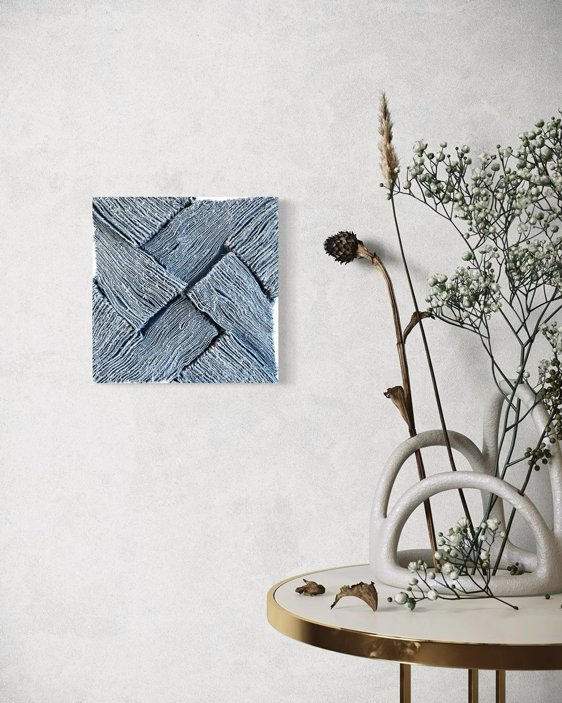 Een witte muur met een kleine vierkante kunstprint met een textuur print, naast een witte vaas met bloemmotieven en gedroogde bloemen op een ronde witte tafel met gouden rand.