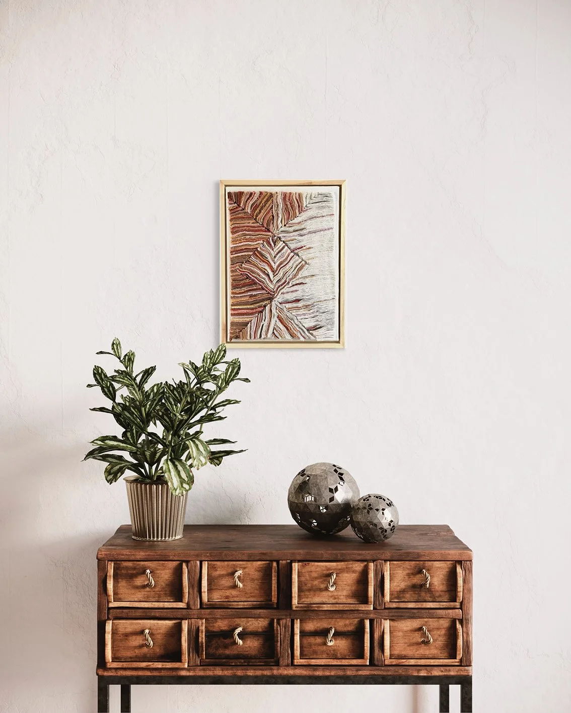 Een houten dressoir met zes kleine laden en touwgreepknoppen, een plantenpot met groene bladeren, en twee decoratieve metalen bollen, hangend aan een witgerekte muur met een abstract kunstwerk.