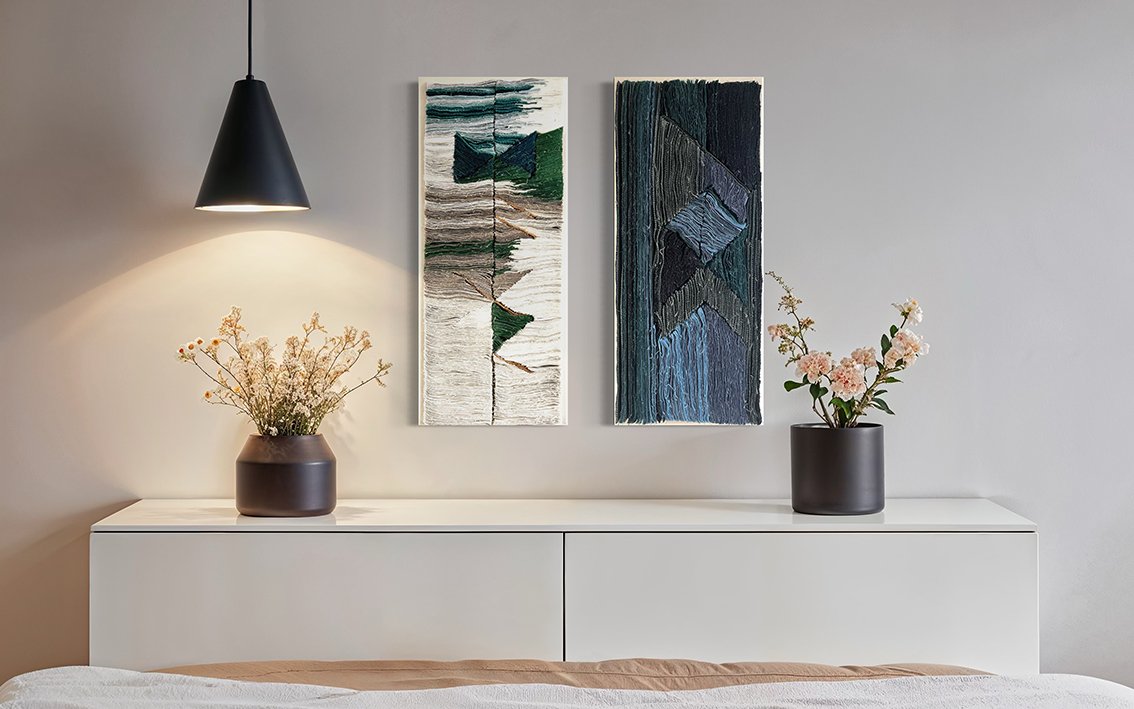 Witte slaapkamer met twee zwarte vazen met bloemen, twee abstracte textiele kunstwerken aan de muur, en een witte kast onder de kunstwerken.