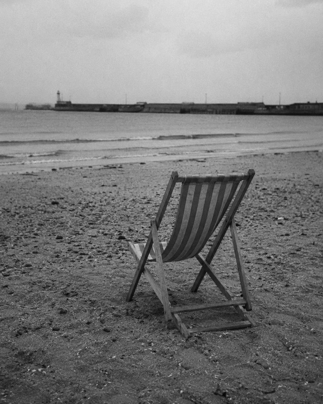 A wooden beach chair facing the ocean on a sandy beach with a distant pier and calm water, black and white photograph.