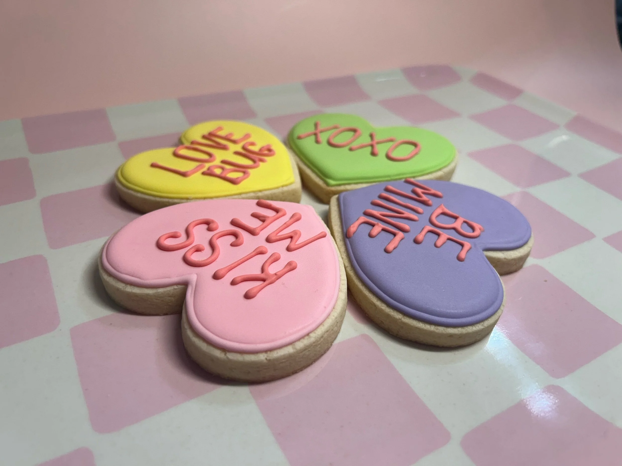 Four heart-shaped cookies with colorful frosting and messages: yellow with 'LOVE LOVE', green with 'XOXO', pink with 'KISS ME', purple with 'BE MINE', on a pink checkered paper.