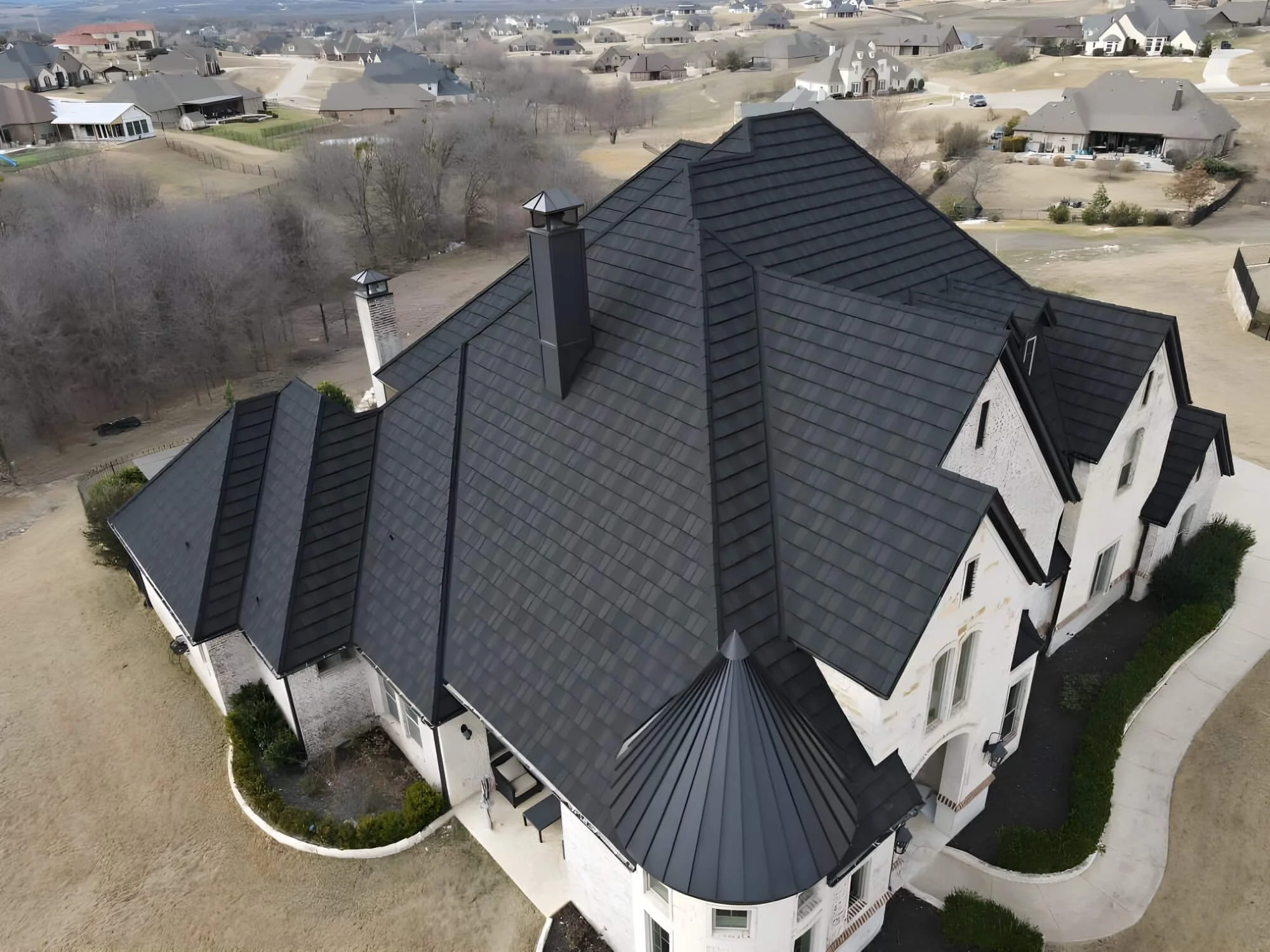 Aerial view of a large white house with a complex dark gray roof, multiple chimneys, and surrounding yard and neighborhood houses.