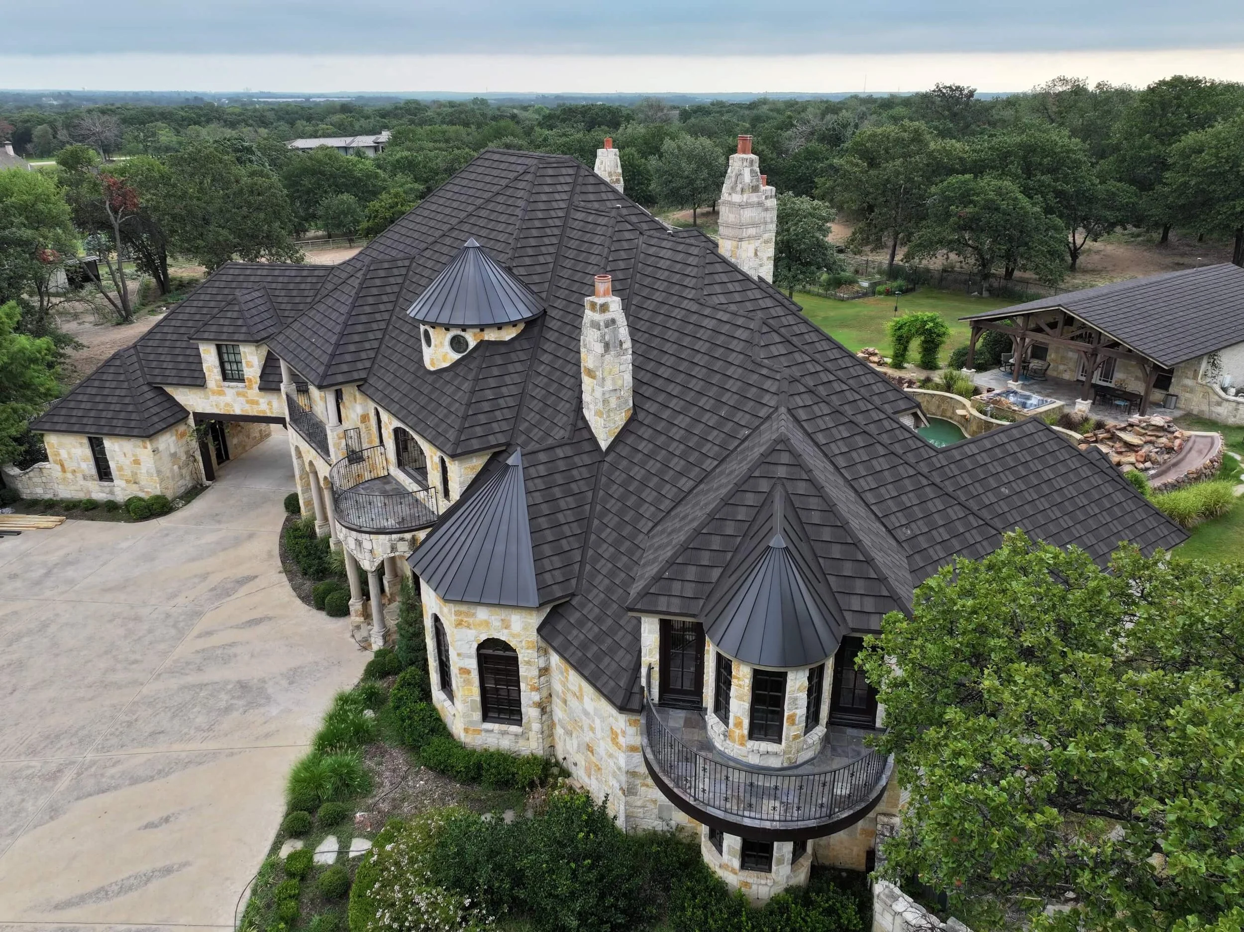 Aerial view of a large, elegant stone mansion with dark tiled roofs, multiple turrets, and tall chimneys, surrounded by lush green trees and landscaped gardens.