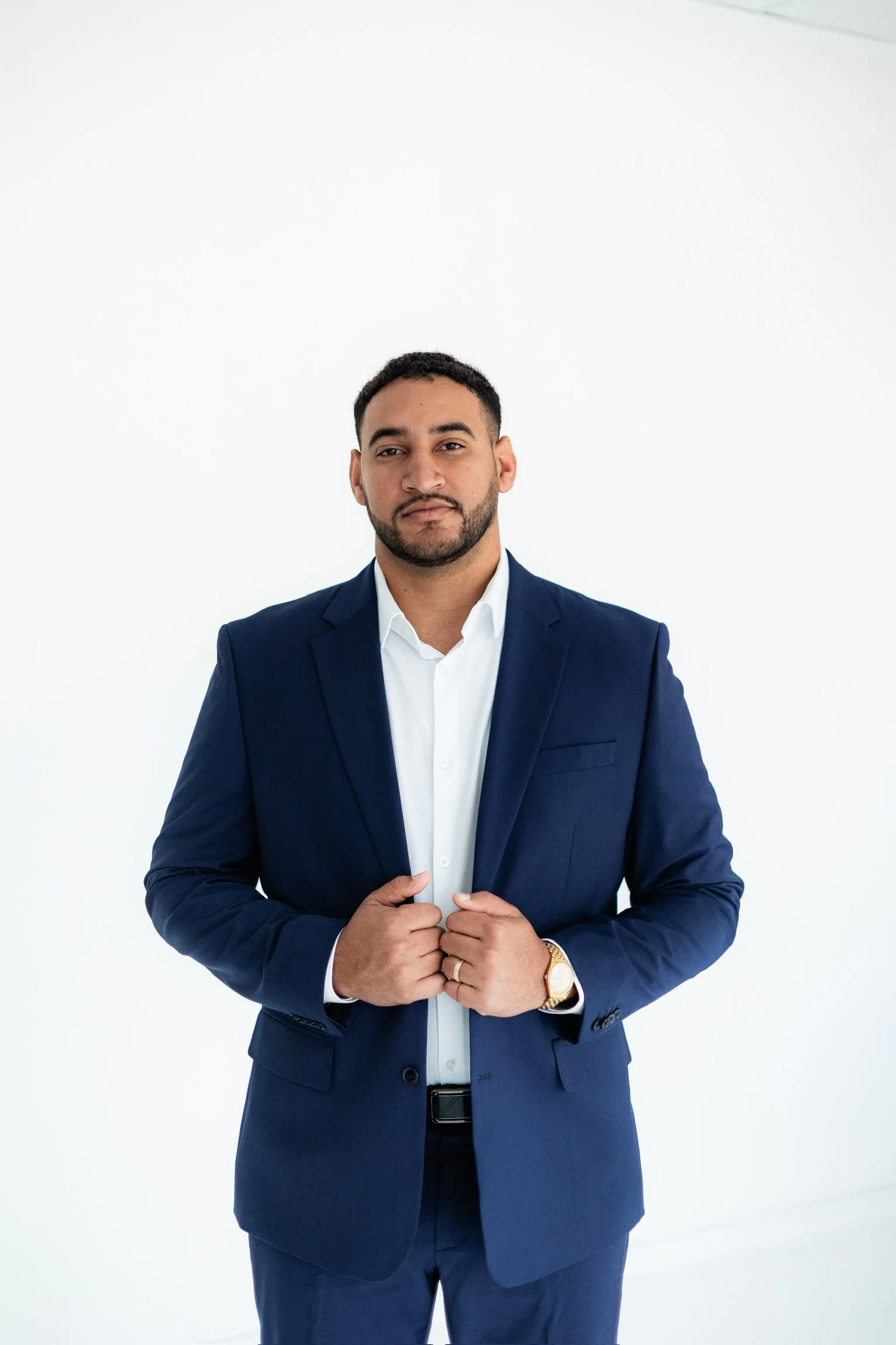 A man in a navy suit and white shirt, standing against a plain white background.