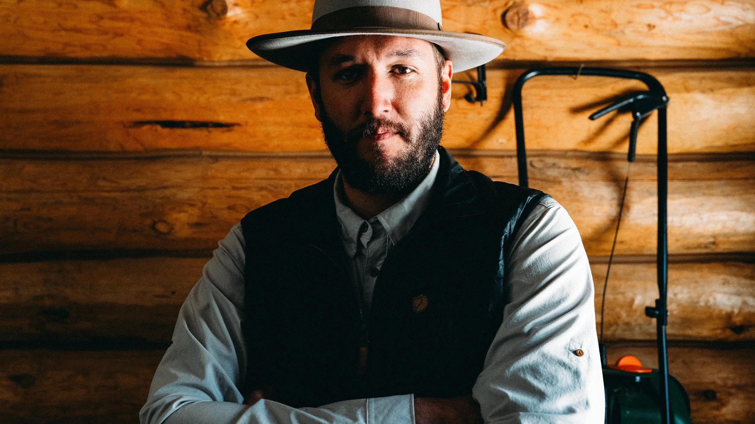 A man with a beard wearing a hat, a light-colored shirt, and a dark vest, posing with arms crossed inside a wooden cabin.
