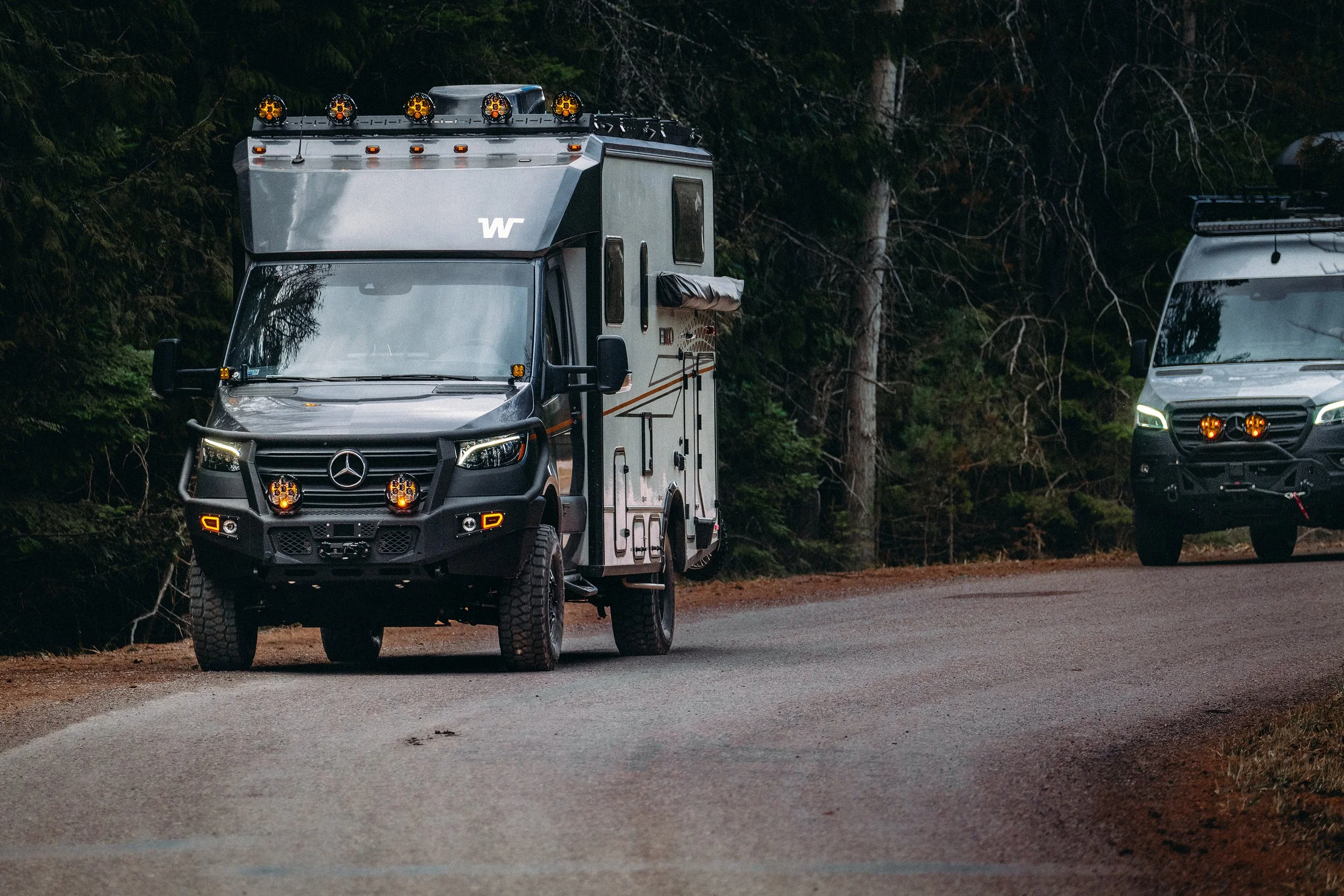 Two dark gray Mercedes-Benz camper vans parked on a gravel forested road, with trees in the background.