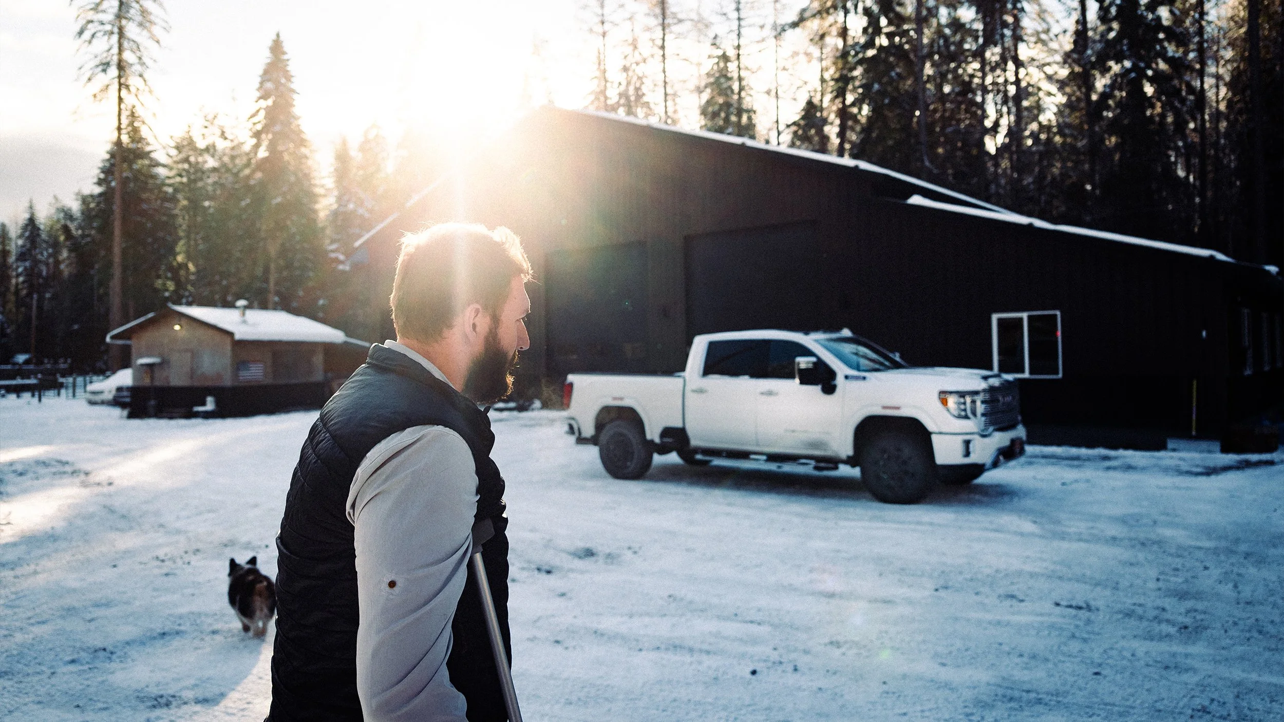 A man with a beard walking on snow with a dog in the background, near a large black building, with sunlight shining through trees in the background.