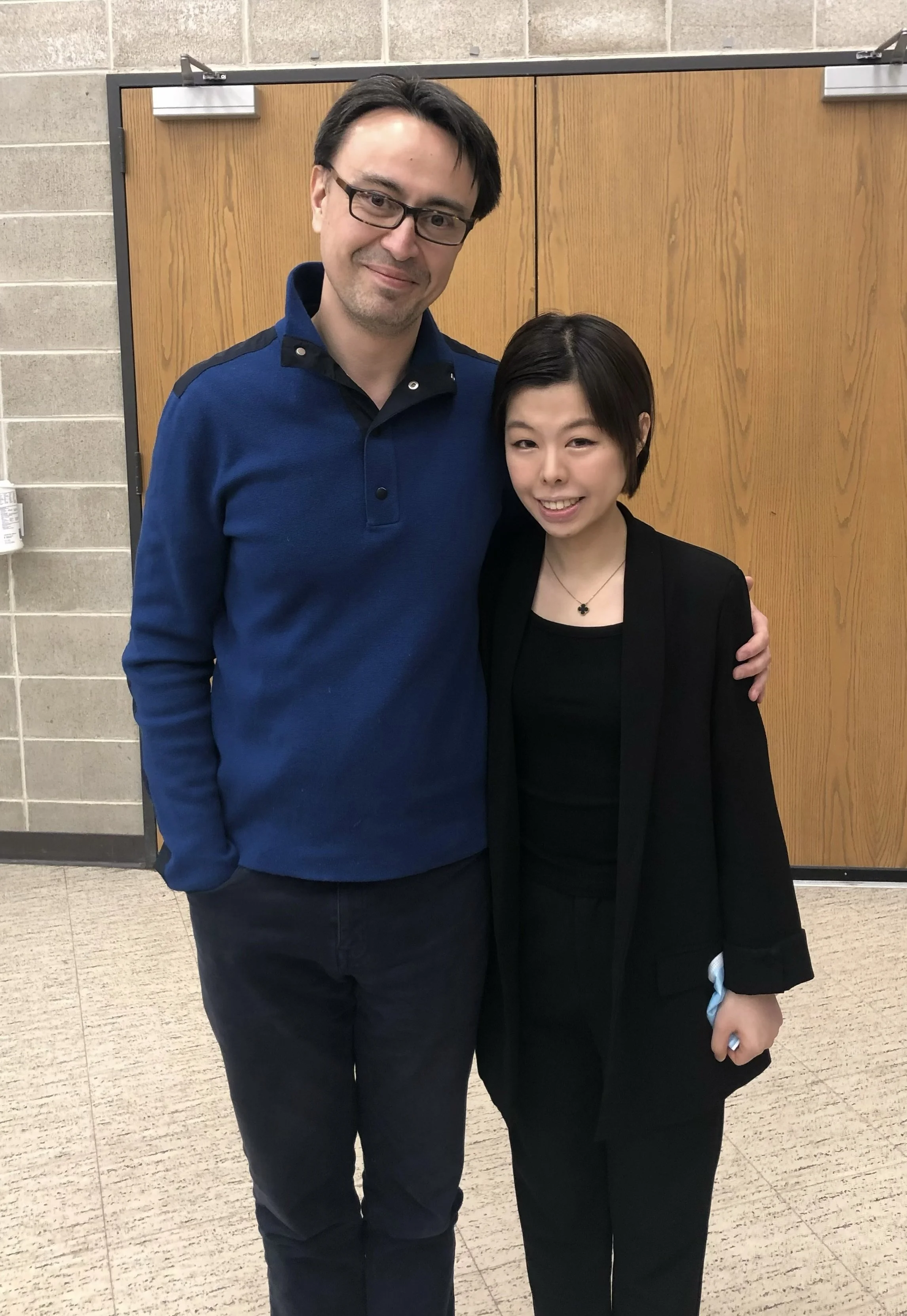 A tall man with glasses and a woman with short dark hair posing together indoors in front of a wooden door.