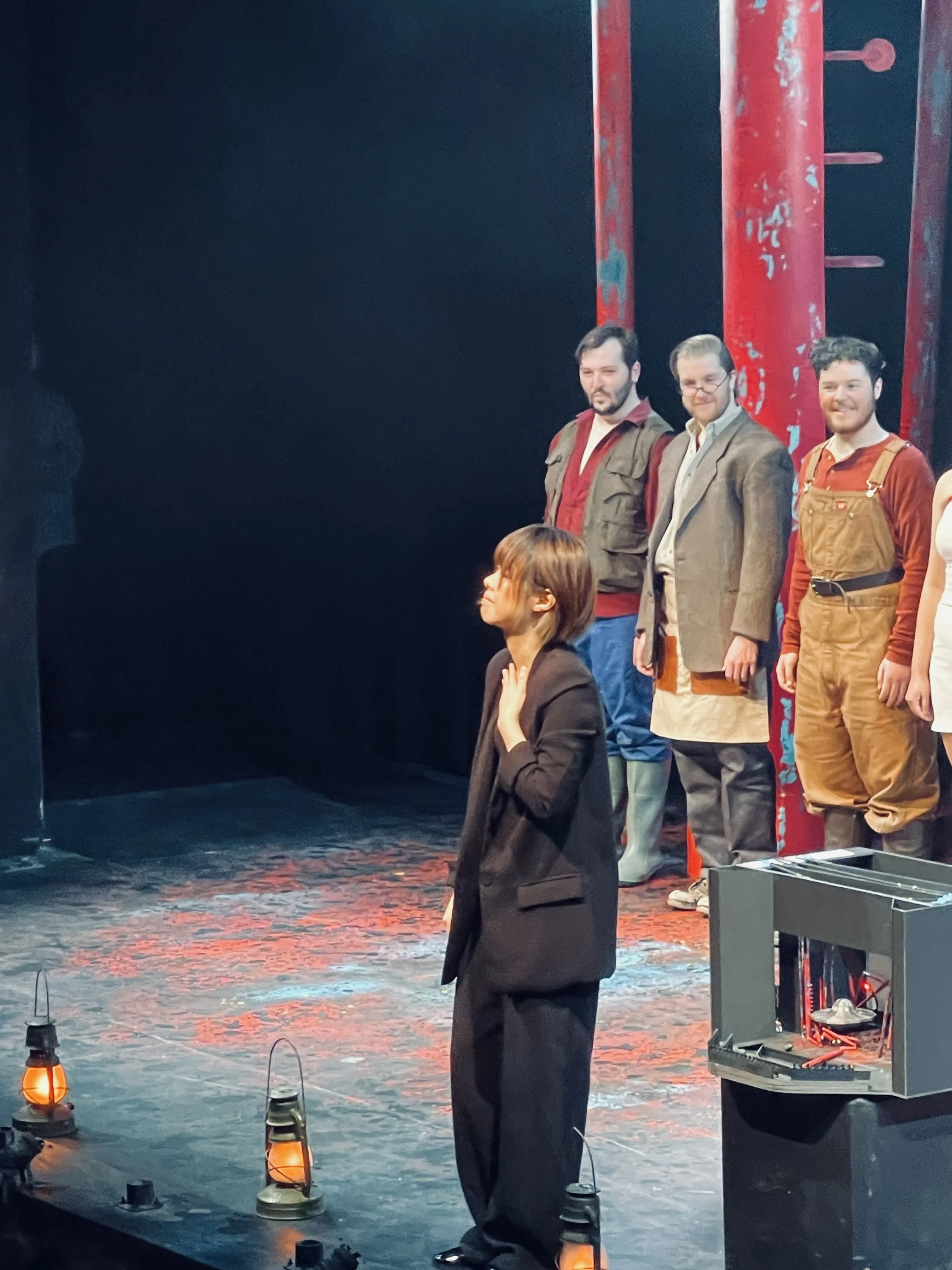 A woman in a black suit stands on stage with her right hand on her neck, facing a group of three men in casual and semi-formal clothing, with a red and black stage backdrop and lanterns on the floor.