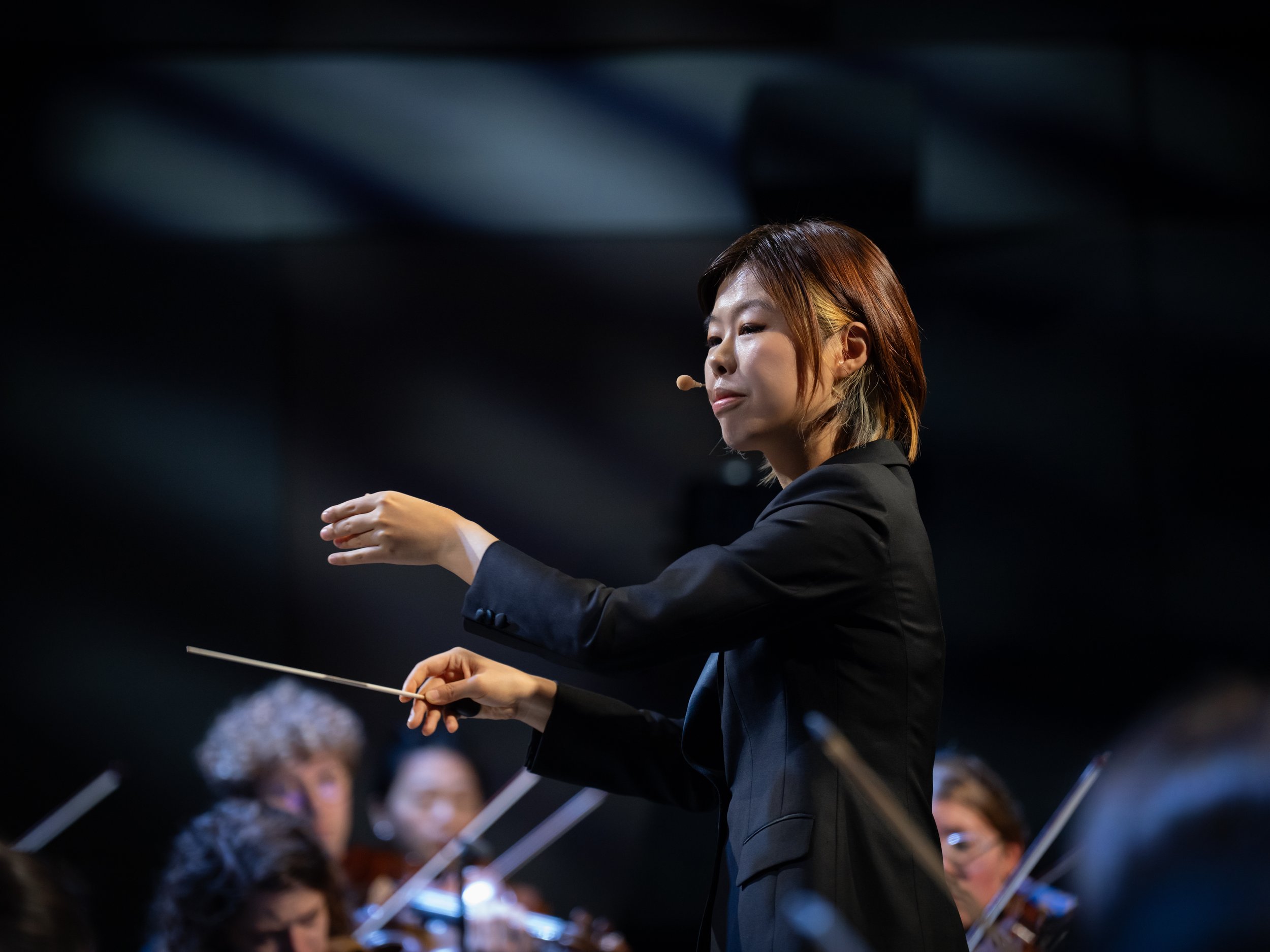A female orchestra conductor directs an orchestra, facing to the right, wearing a black blazer, with a blurred orchestra in background.