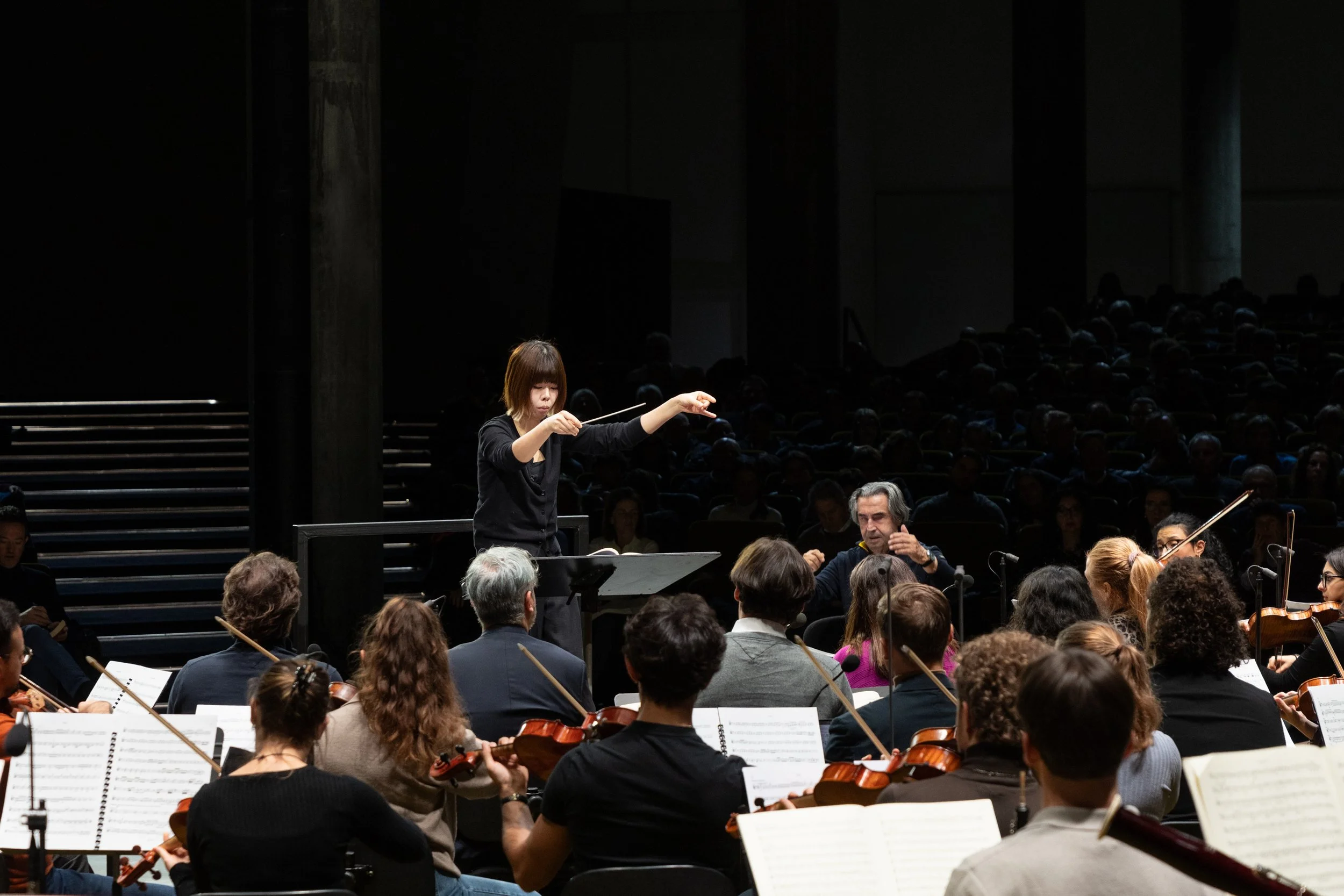 A woman conducts an orchestra during a performance, with an audience in the dark background.