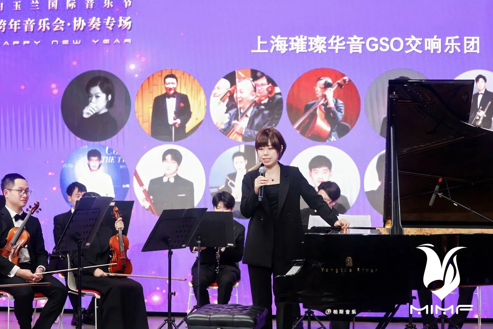 A woman in a black suit and short hairstyle speaking into a microphone on stage with a grand piano and orchestra, during a performance or event. The background has a purple screen with photographs of musicians and text in Chinese.