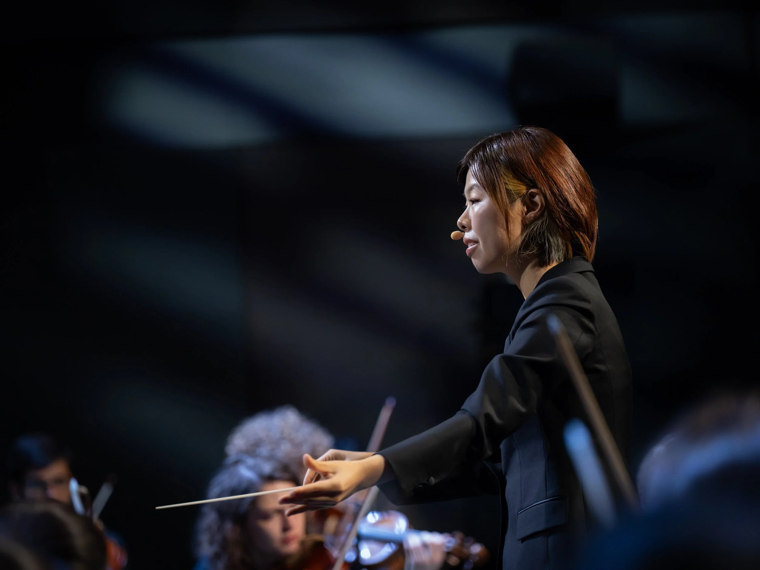 Conductor leading an orchestra, facing sideways with eyes closed, wearing a black suit, during a performance.