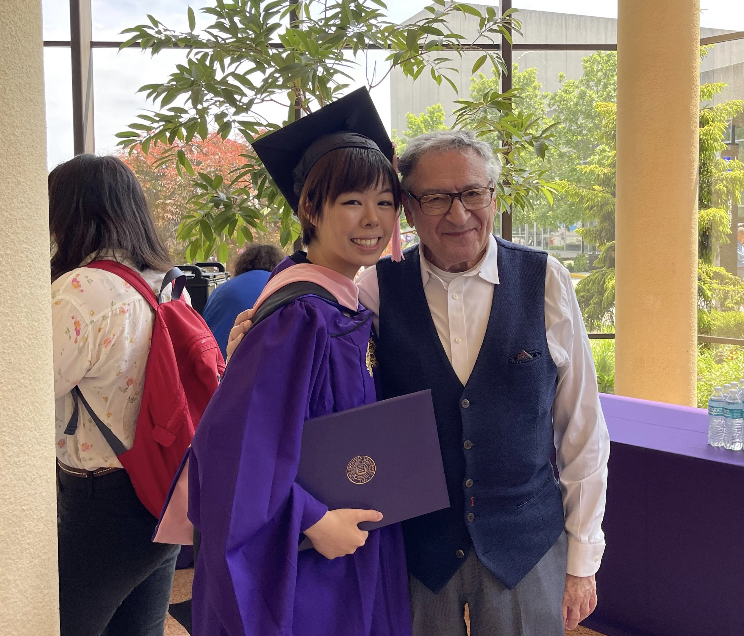 A young woman in a purple graduation gown and cap holding a diploma, standing next to an older man in a white shirt and vest, at a graduation ceremony indoors near large windows with greenery outside.