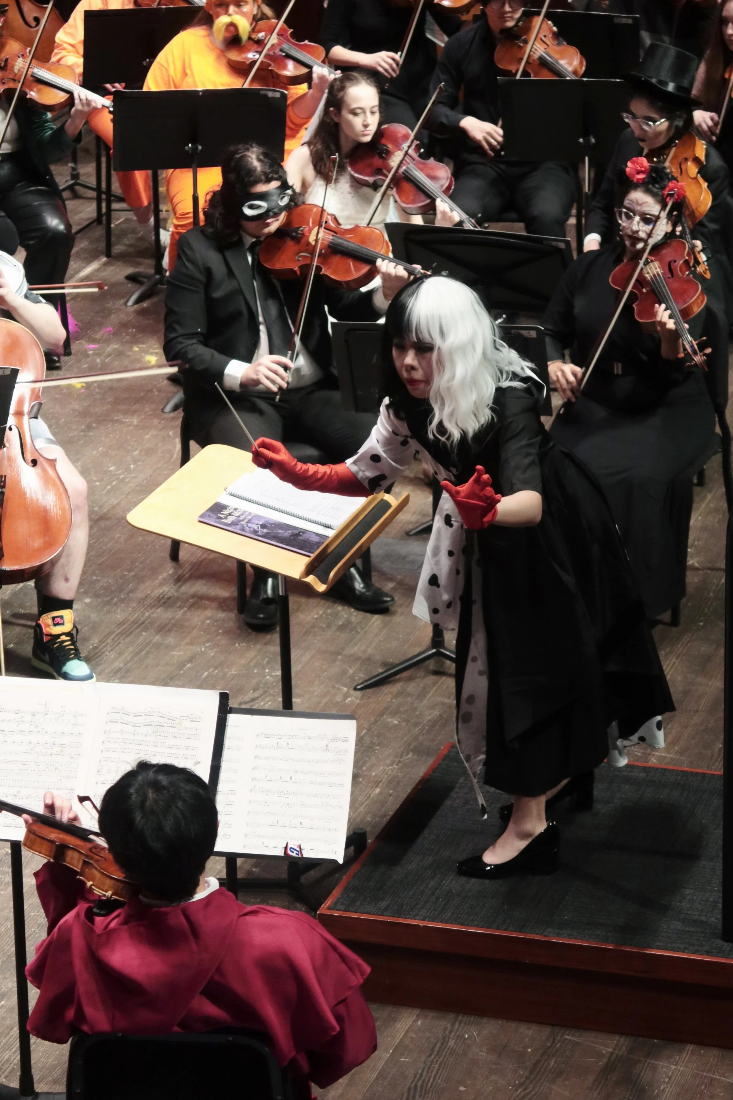 Orchestra performing on stage, with an actor conductor wearing a white wig, polka-dot dress, red gloves, and in-character costume, conducting the musicians.