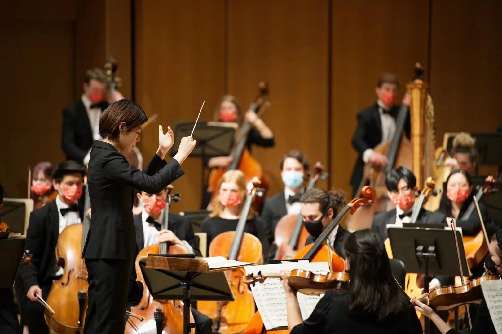 Orchestra in performance with conductor and musicians wearing masks, playing string instruments, in concert hall.