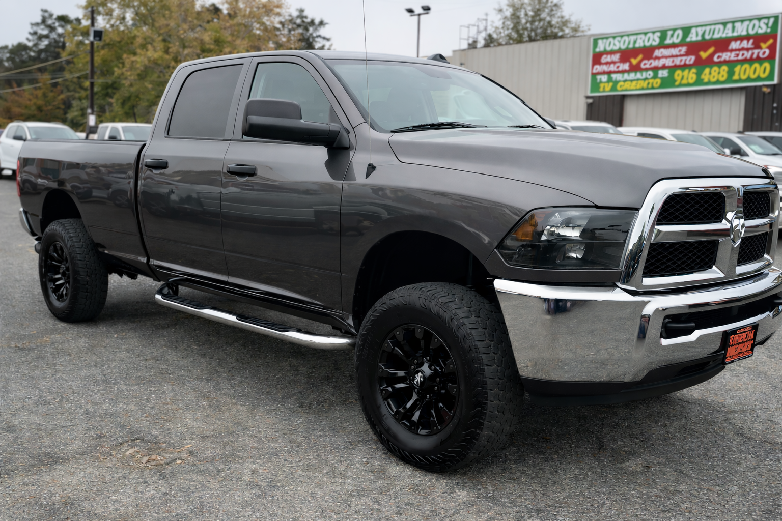 Gray Dodge Ram pickup truck parked on a gravel lot, dealership sign in the background with text in Spanish.