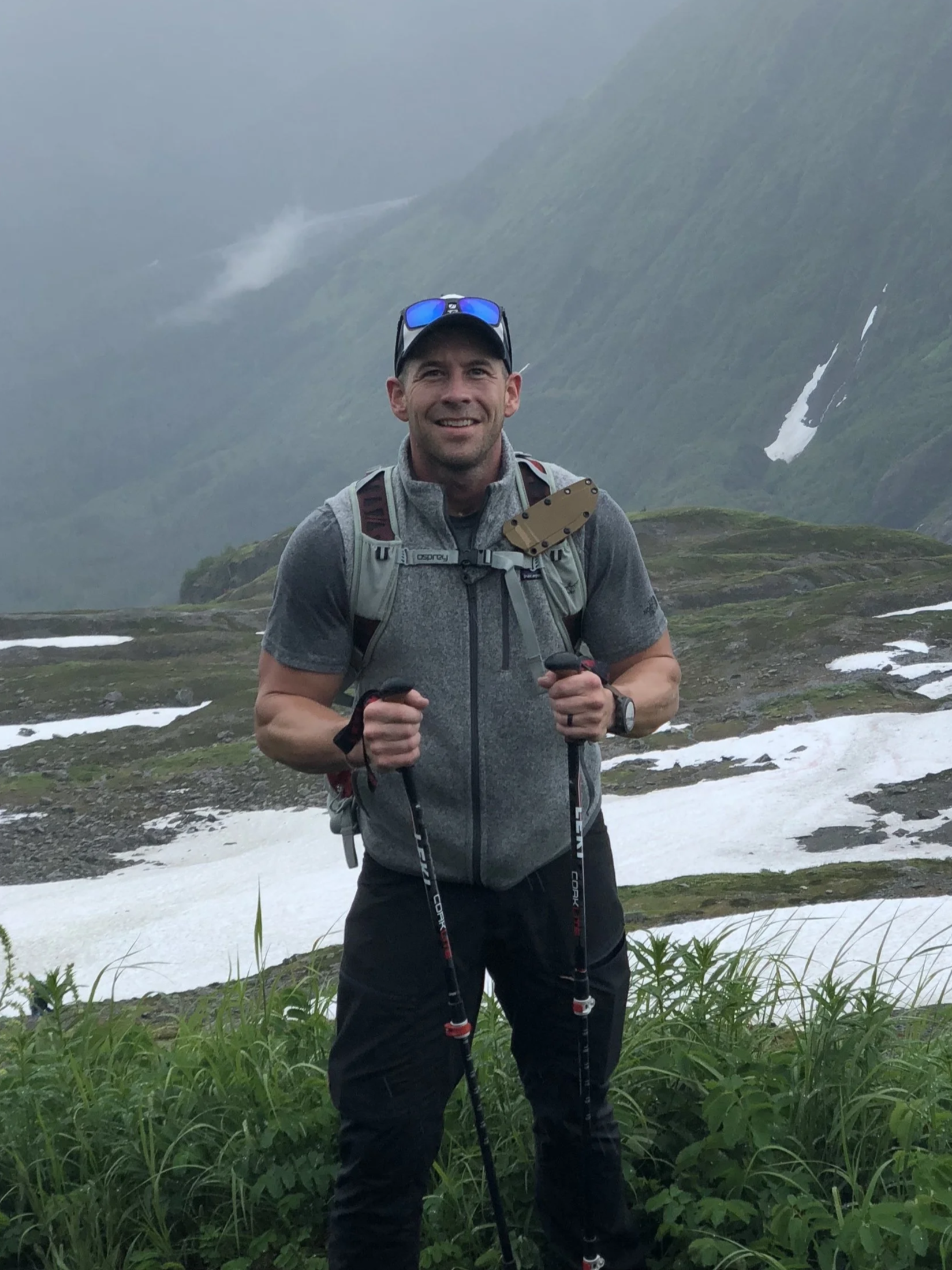 A man hiking outdoors in a mountainous area with snow patches, wearing a gray vest, black pants, a watch, sunglasses on his head, and holding hiking poles.