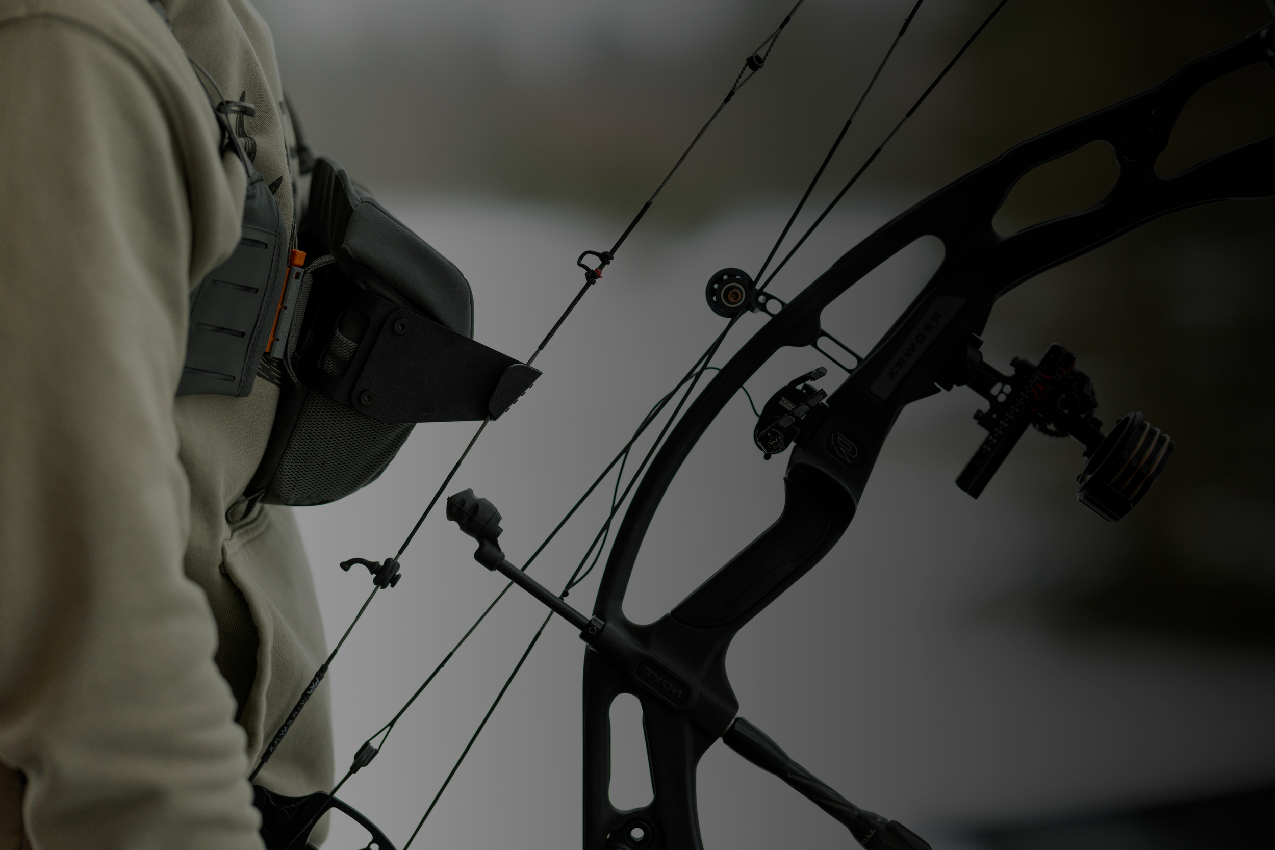 Close-up of a person's upper body holding a modern compound bow while carrying a quiver with arrows on their chest.