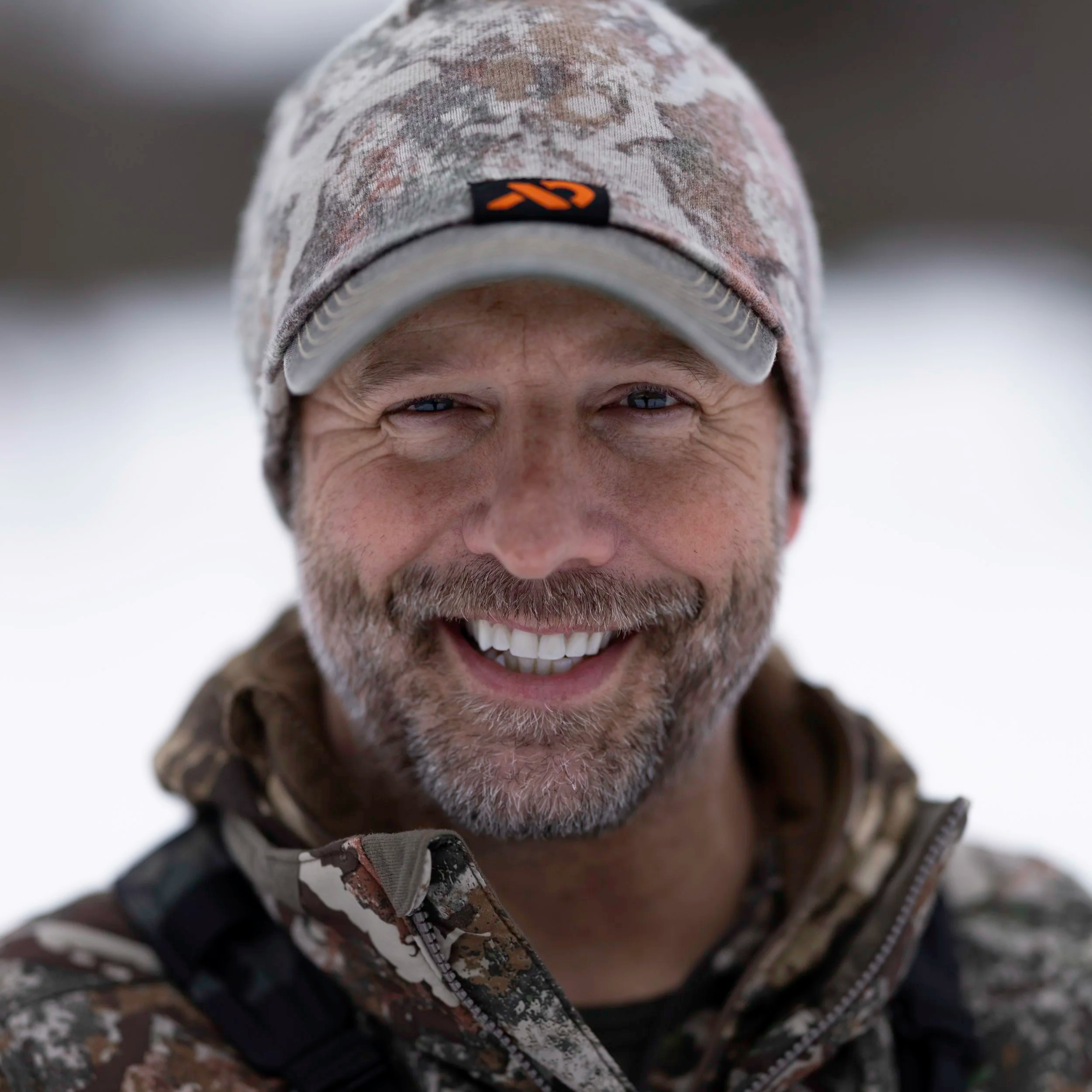 Close-up of a smiling man wearing a camouflage jacket and matching camouflage cap in a snowy outdoor setting.