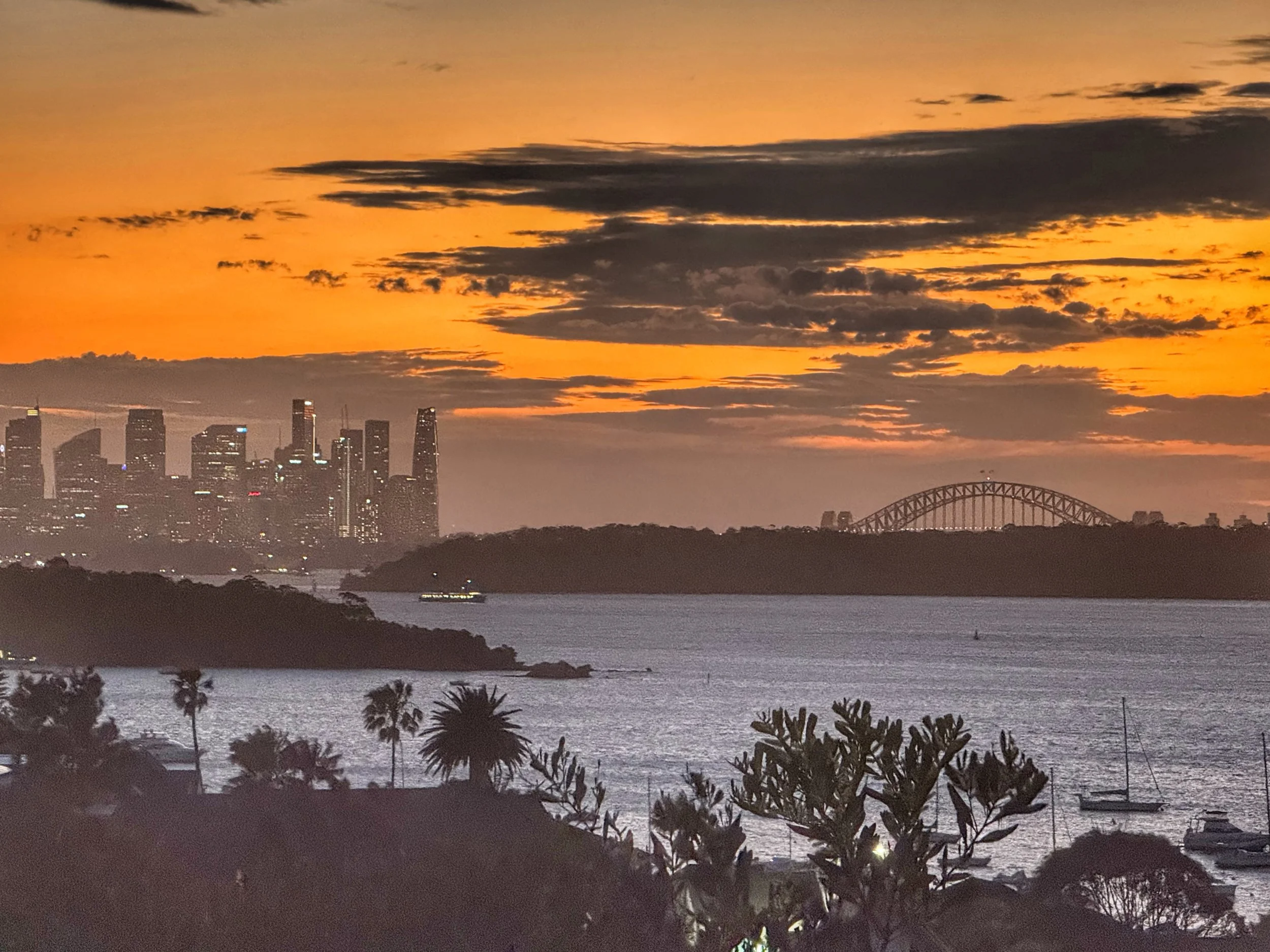Atardecer sobre la ciudad de Sydney, con el horizonte de rascacielos y el puente de la Bahía de Sydney al fondo, junto a un paisaje con vegetación y barcos en el agua.