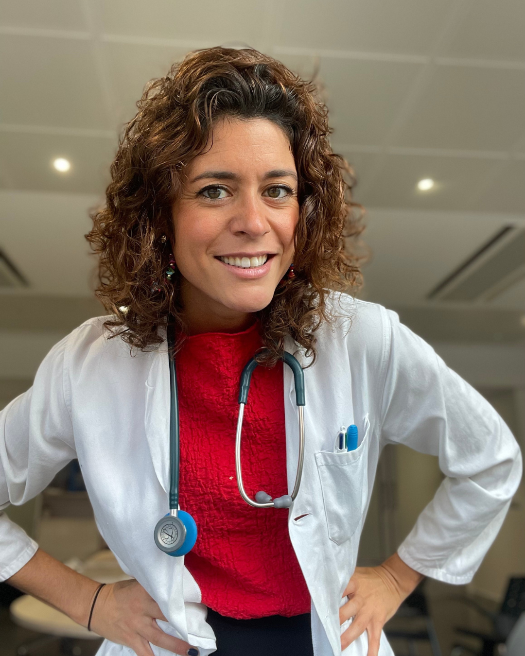 Médica mujer con estetoscopio en el cuello, sonrisa, fondo de consultorio.