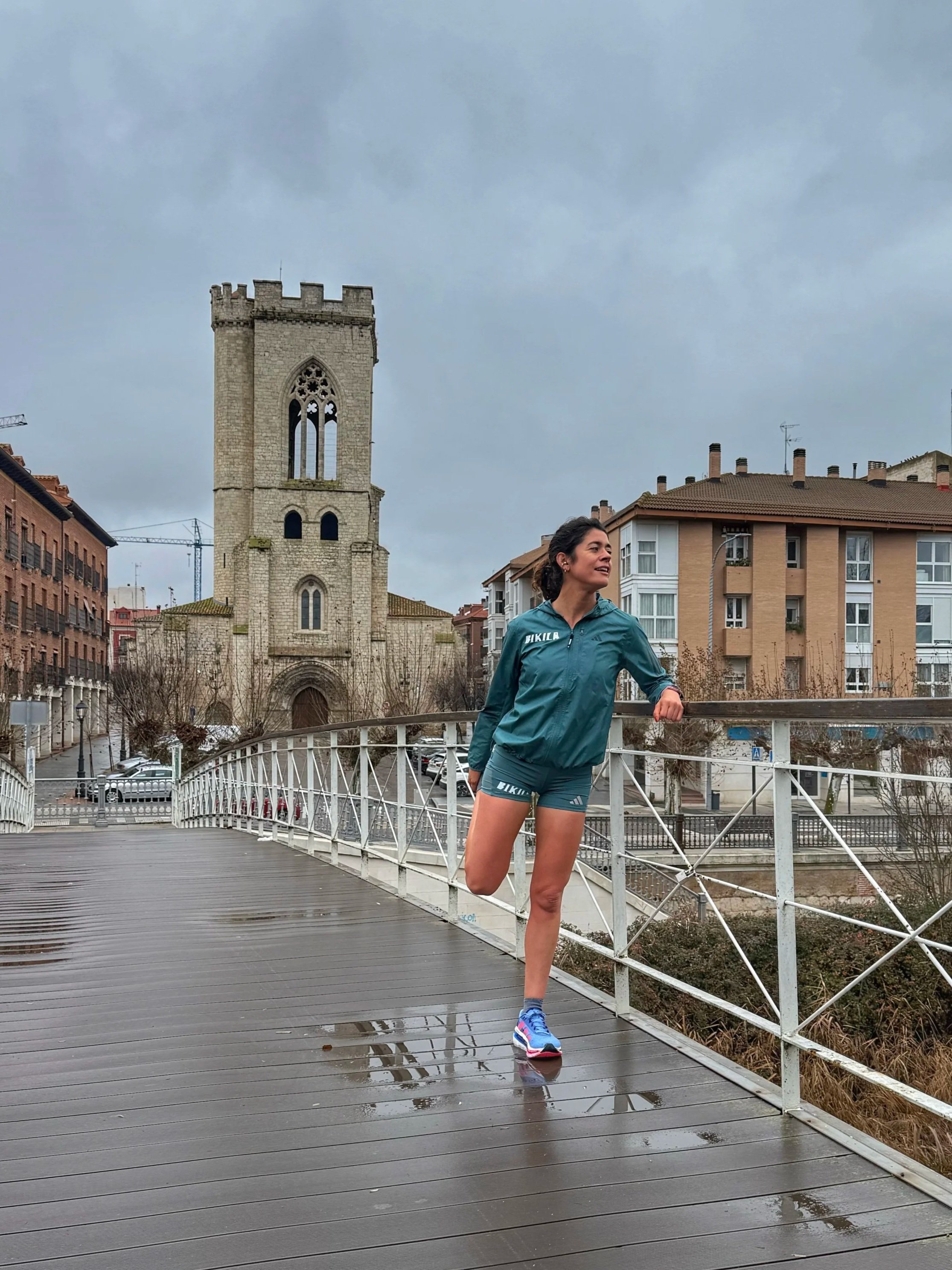 Una mujer corriendo en un puente de madera en un día nublado, con una iglesia y edificios residenciales de fondo.