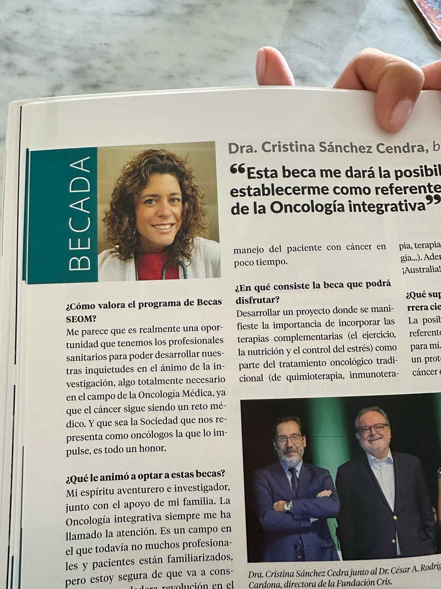 Página de revista con una entrevista a la Dra. Cristina Sánchez Cendra, con una fotografía de ella y otra de ella con un hombre. Texto en español que menciona una beca y su interés en la oncología integrativa.