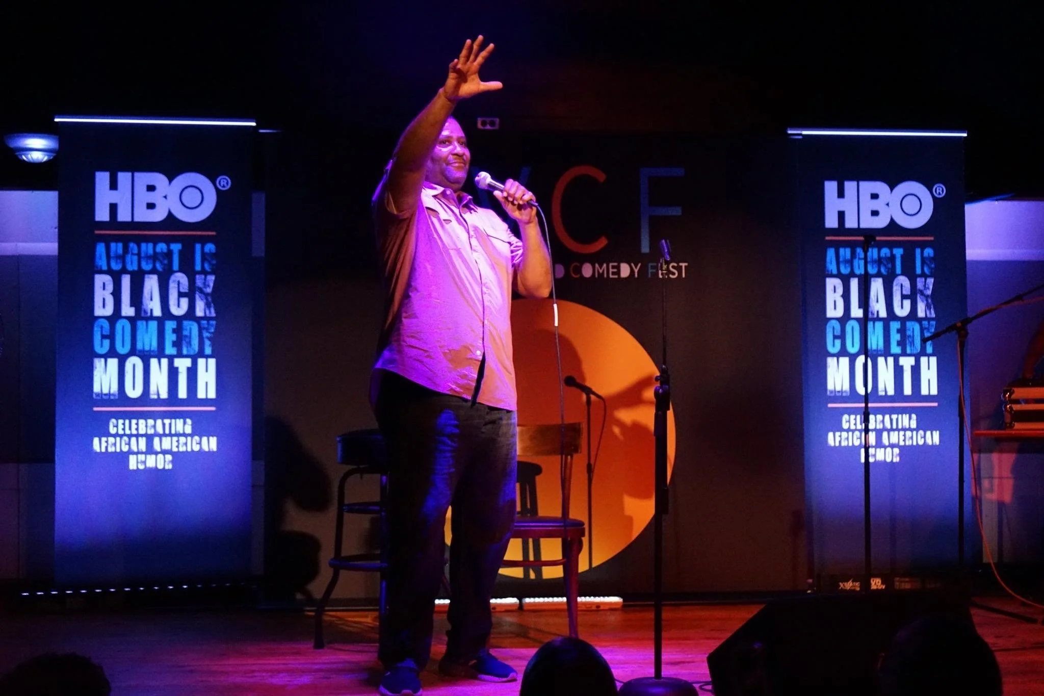 Comedian performing on stage at an event celebrating Black American Month, with HBO banners on either side.