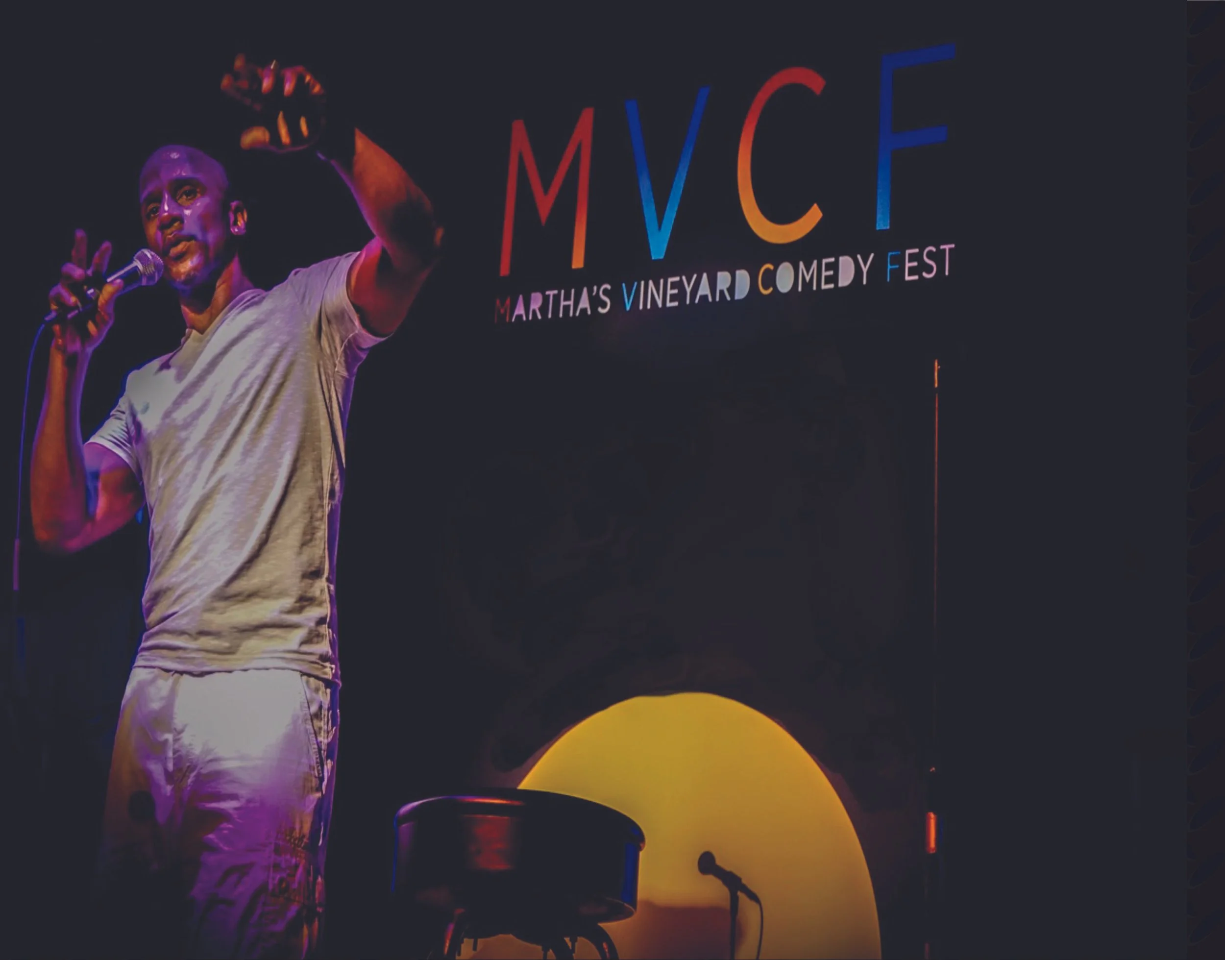A man stands on stage holding a microphone in front of a colorful sign that reads 'MVCF' for Martha's Vineyard Comedy Fest, with a dark background and stage lighting.