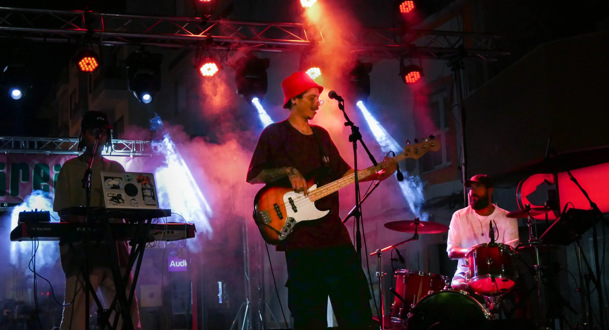 Musiker auf einer Bühne, die mit buntem Lichter und Rauch für einen Live-Auftritt beleuchtet wird: eine Person spielt Bass, eine andere an elektronischen Geräten, eine weitere am Schlagzeug.