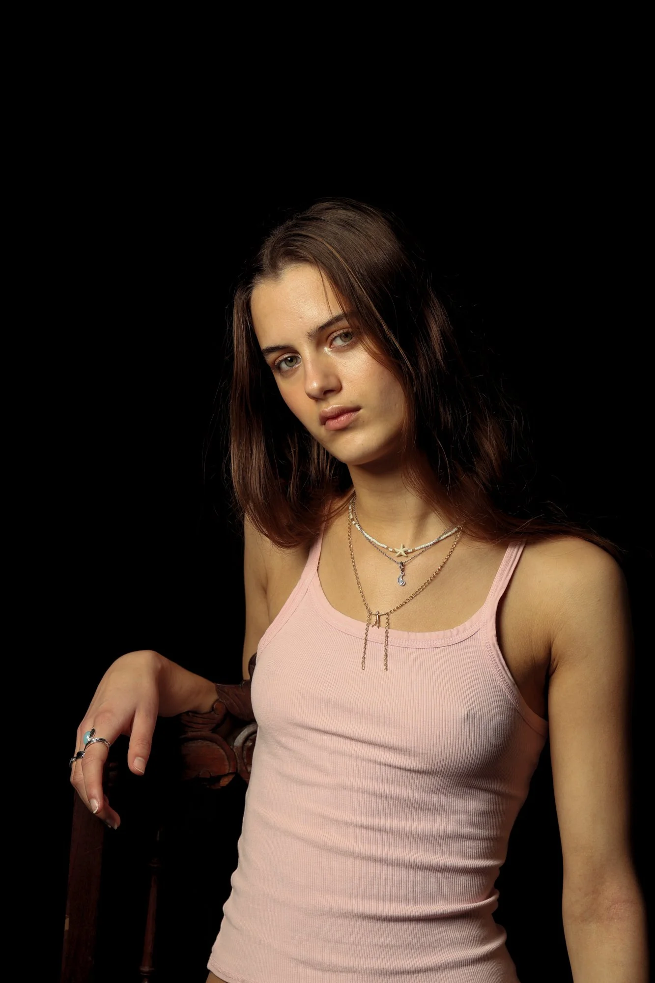 Young woman with brown hair wearing a pink tank top and layered necklaces, leaning on a wooden chair against a black background.