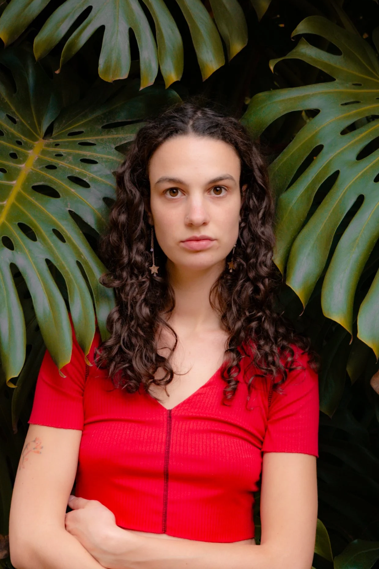 Junge Frau mit lockigem Haar in rotem Oberteil vor großen, grünen Monstera-Blättern.
