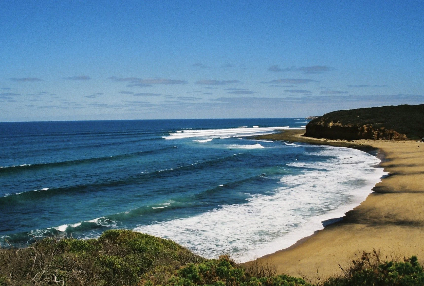 Ein Strand mit goldgelbem Sand, umgeben von grünen Büschen und einem Kliff im Hintergrund, unter einem blauen Himmel mit wenigen Wolken, Wellen rollen ans Ufer.