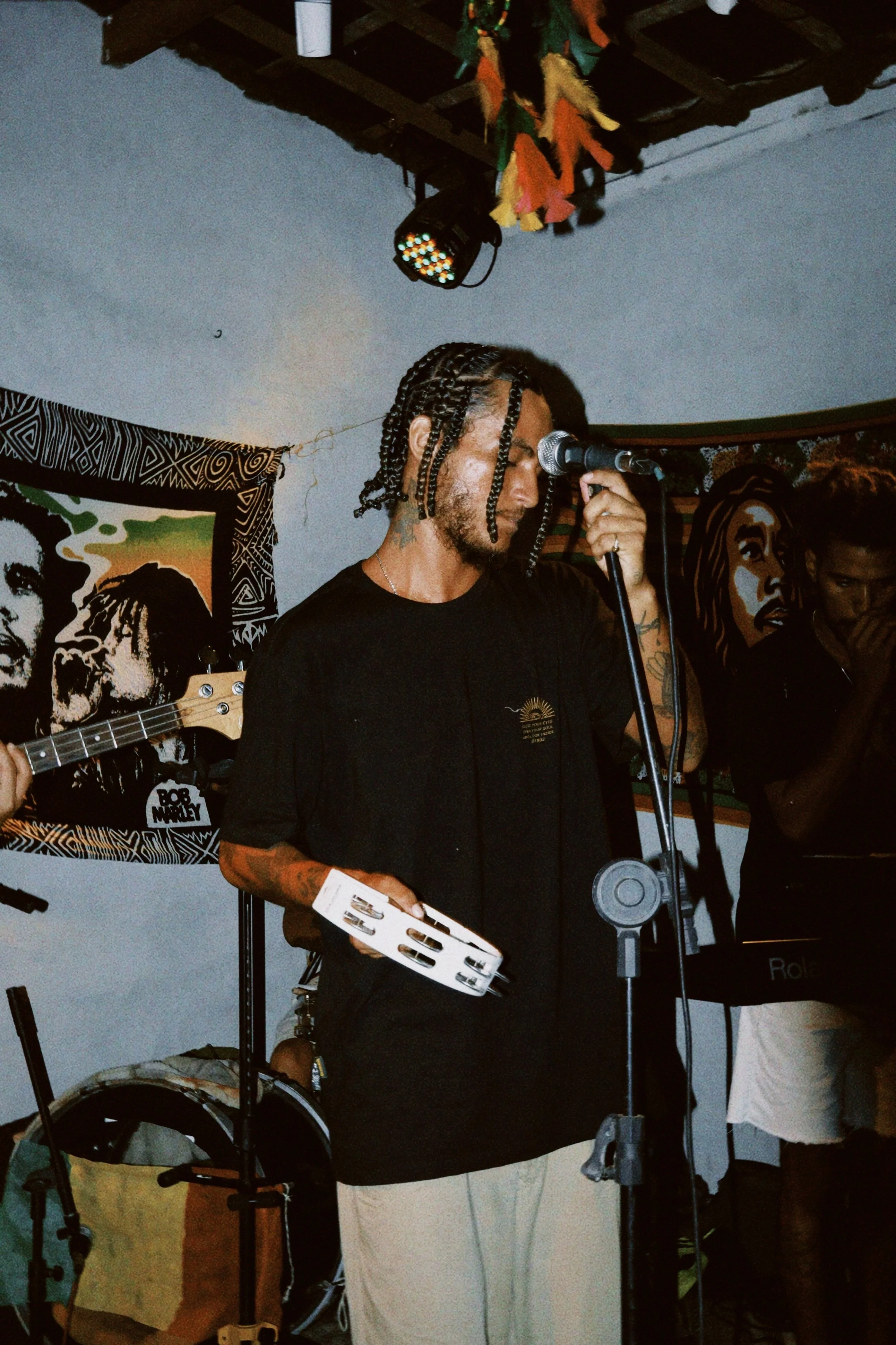 Mann mit dreadlocks hält ein Tamburin und singt in ein Mikrofon, im Hintergrund sind Musikposter und andere Musiker auf einer Bühne.