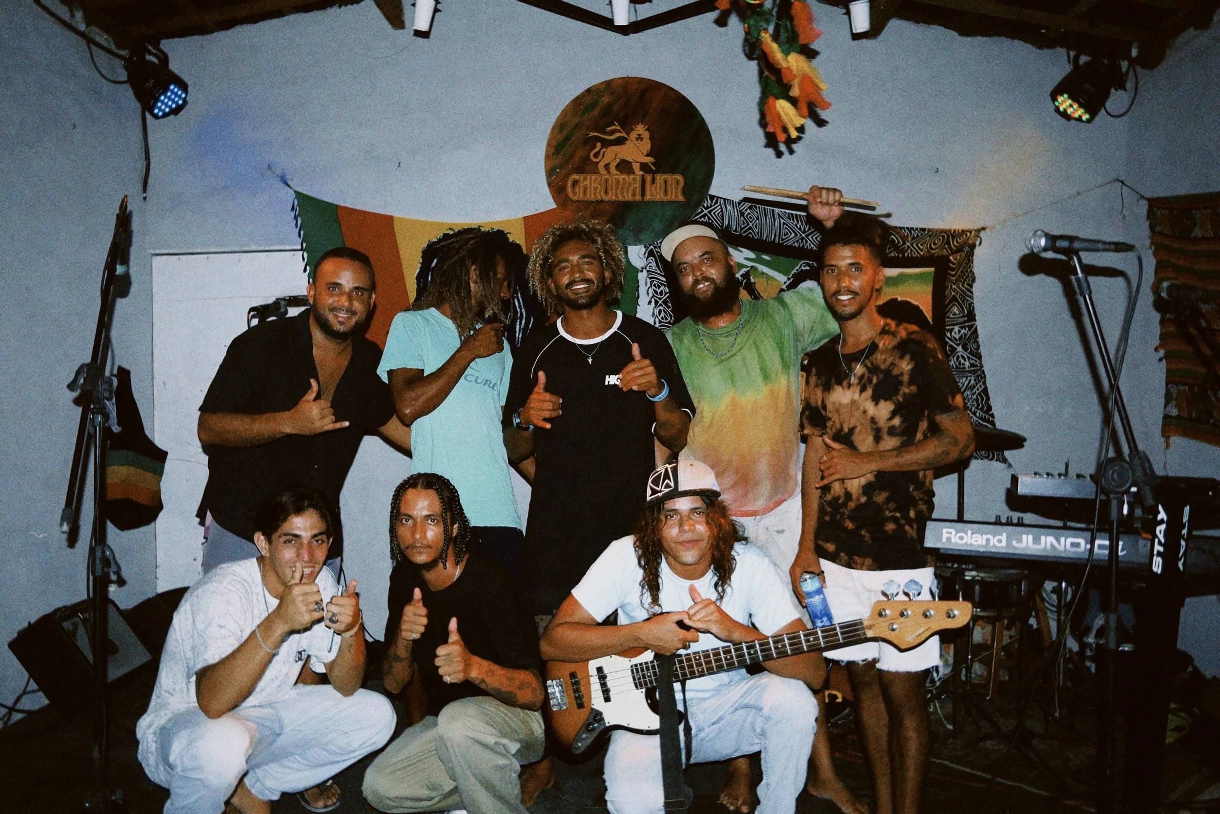 Gruppe von Menschen bei einem Musik-Event, einige mit Musikinstrumenten, im Hintergrund bunte Dekoration, Dachlampe, Mikrofon, Keyboard und Lautsprecher.