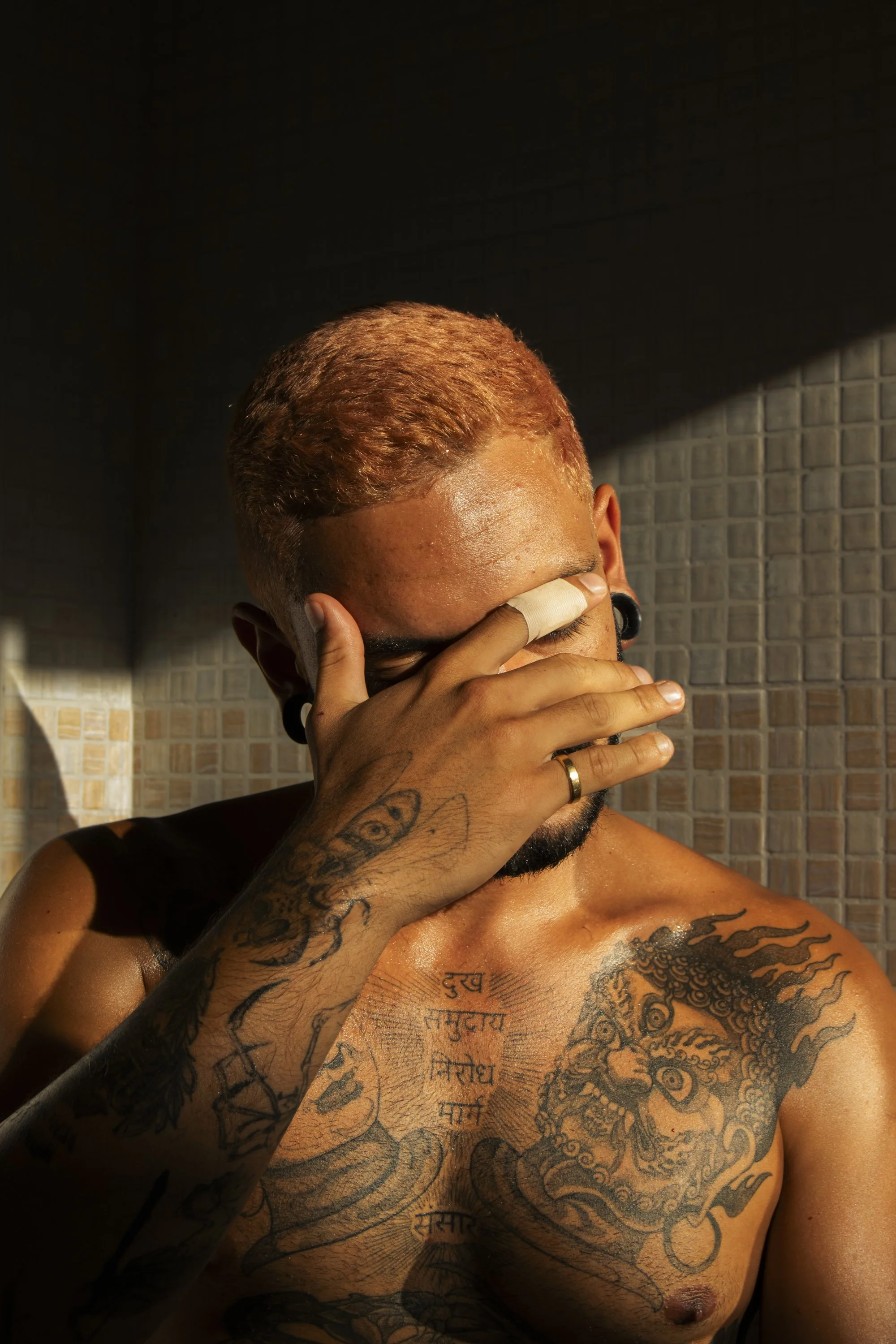 A shirtless man with tattoos covering his chest and arm, holding his face with his hand, in a dimly lit room with a tiled wall background.
