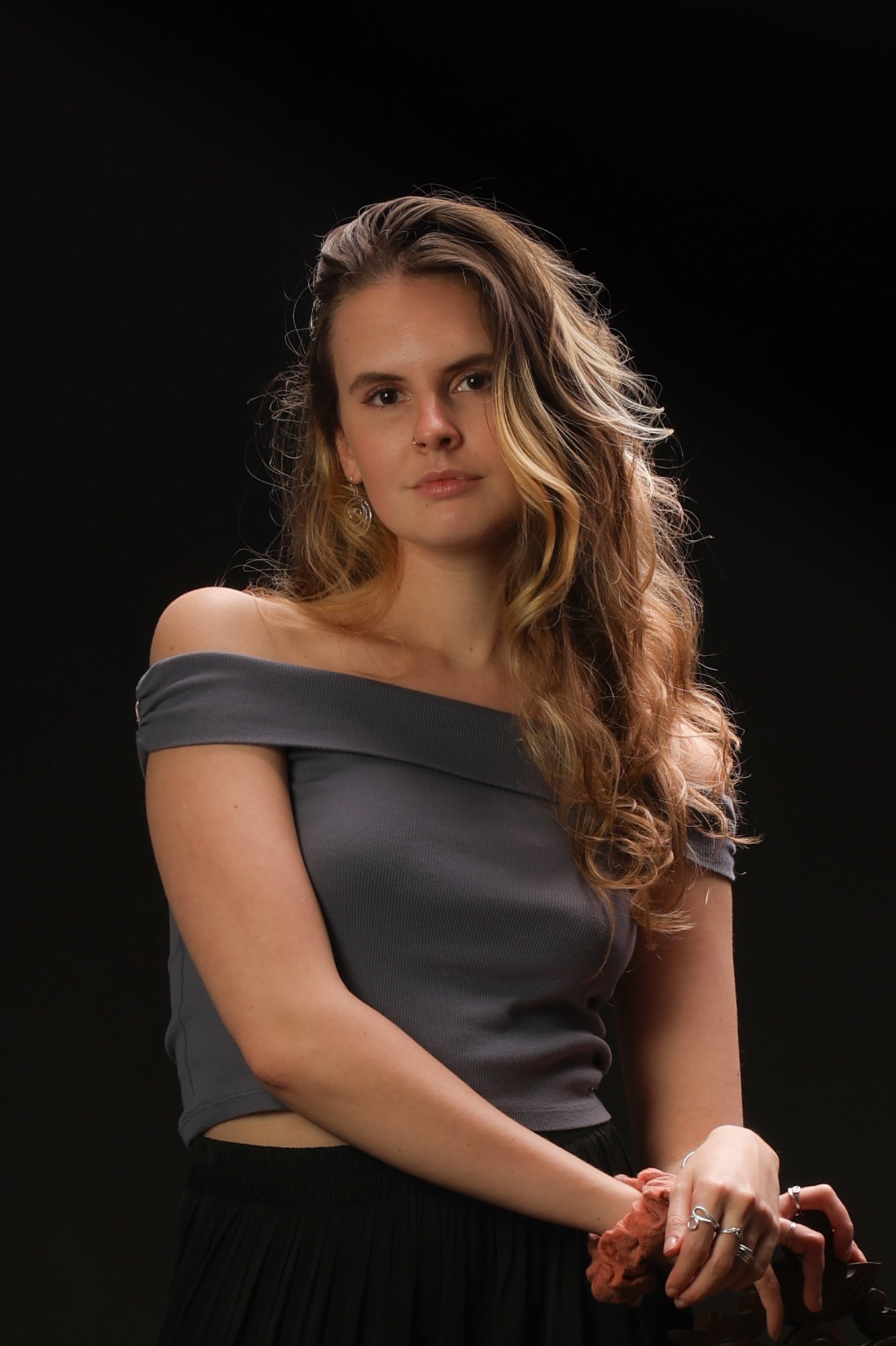 Portrait of a young woman with wavy hair, wearing an off-the-shoulder gray top, standing against a dark background.