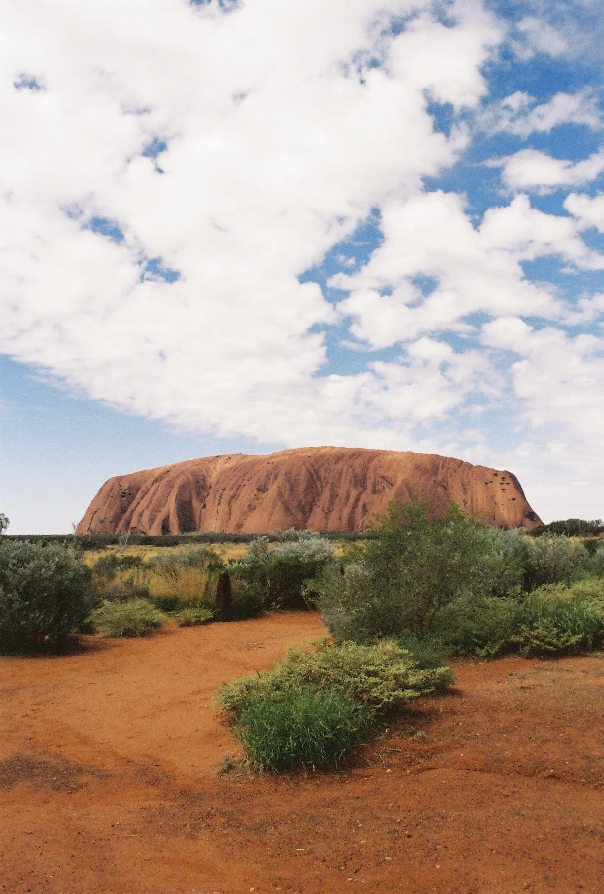 Der Ayers Rock (Uluru) im Outback, Australien, bei sonnigem Himmel mit Wolken, umgeben von Buschland und rotem Sandboden.