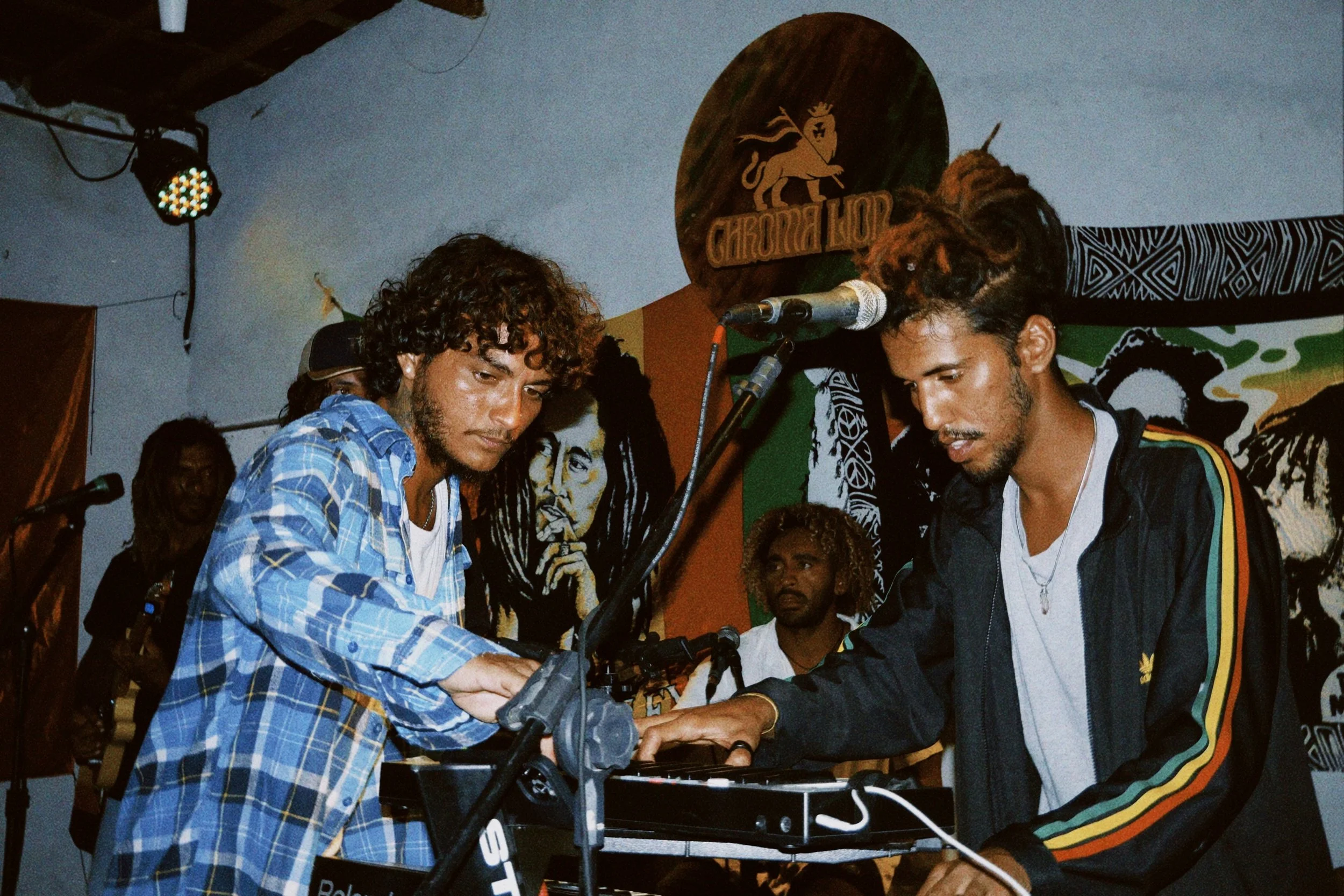 Zwei junge Männer spielen an einem Keyboard bei einem Live-Musikauftritt in einem Raum mit bunten Wandbildern und einem an einer Wand befestigten Logo, im Hintergrund eine weitere Person mit Gitarre.