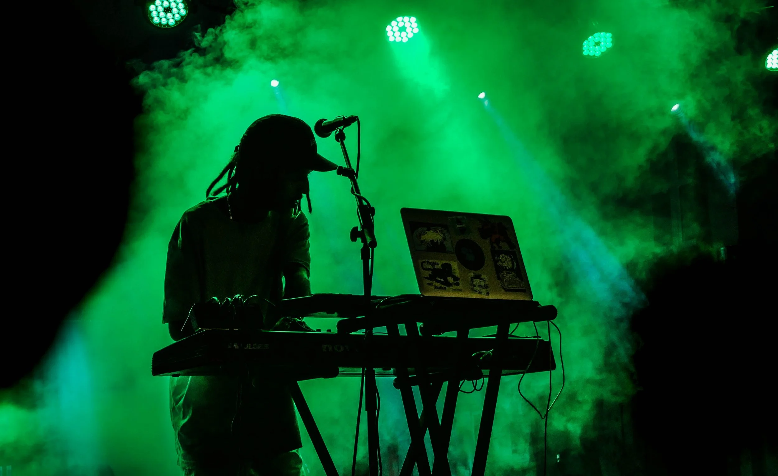 Silhouette eines DJs mit Kopfhörern, der auf einem DJ-Controller und einem Laptop an einem mit grünen Lichtern beleuchteten Veranstaltungsort spielt.