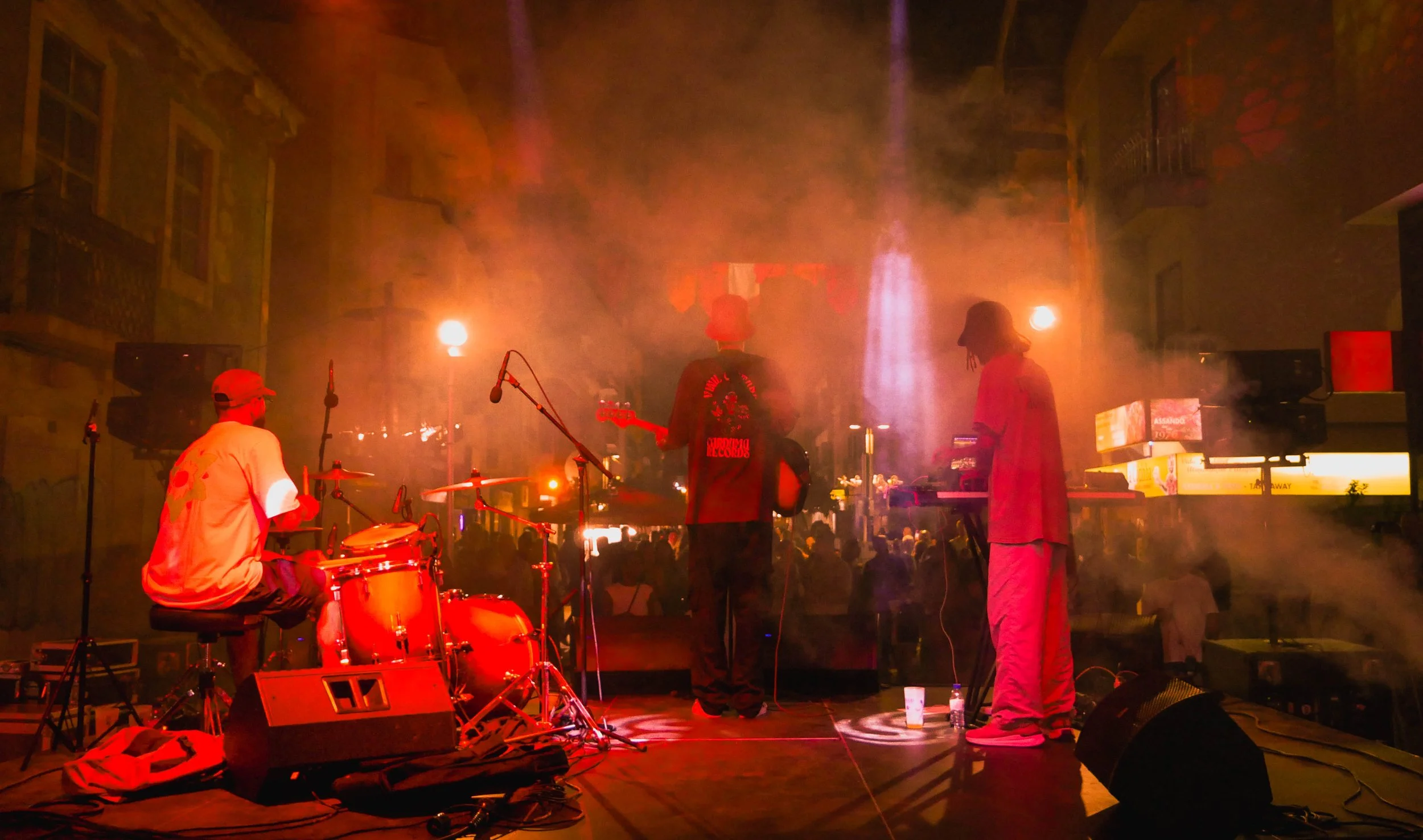 Musiker auf einer Bühne während eines Konzerts, mit Rauch und Licht, in einer urbanen Umgebung.
