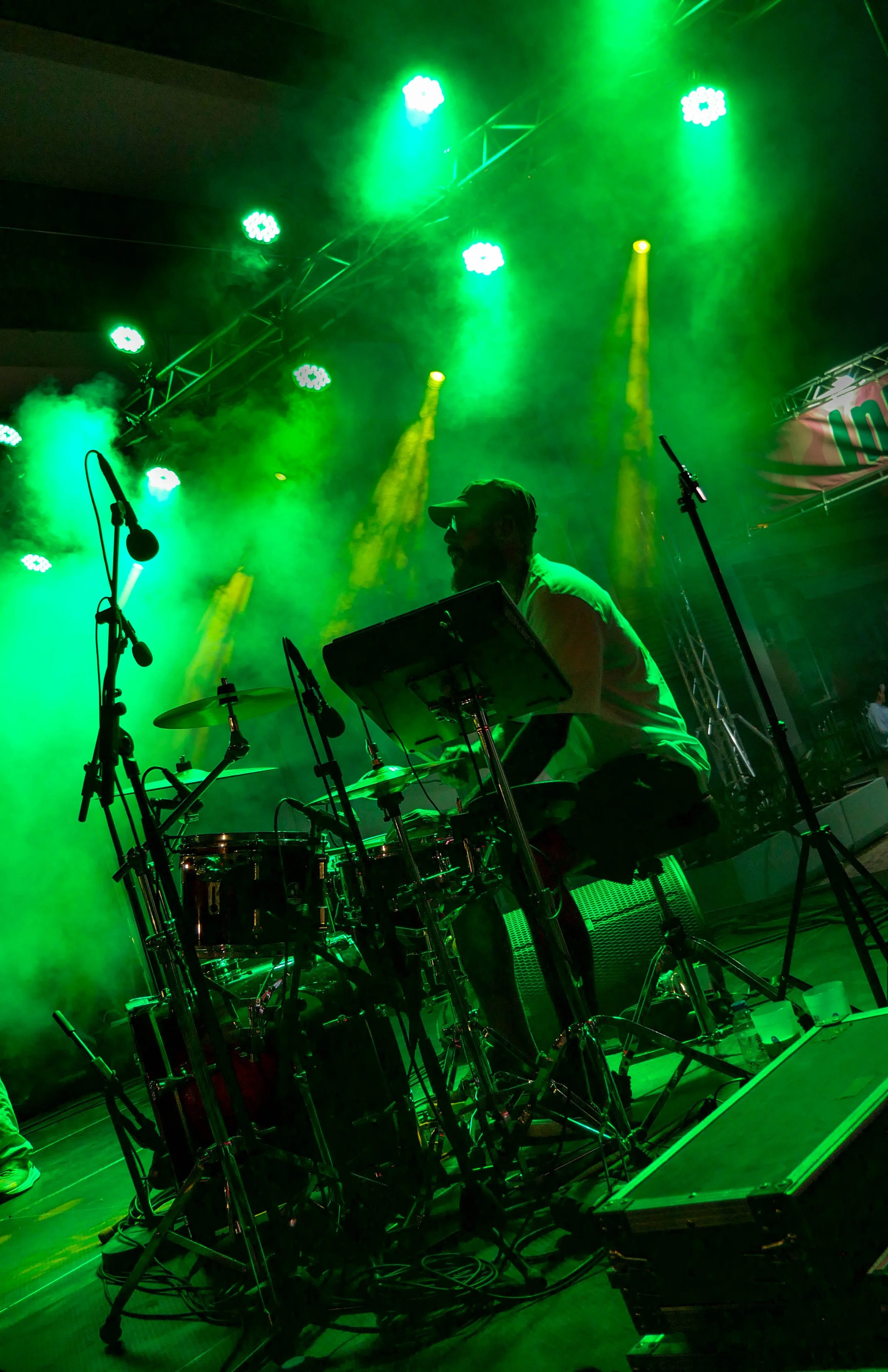 Musiker sitzt hinter einem Schlagzeug auf einer Bühne, umgeben von grünen Lichteffekten und Nebel, mit Mikrofonen und Musikzubehör.