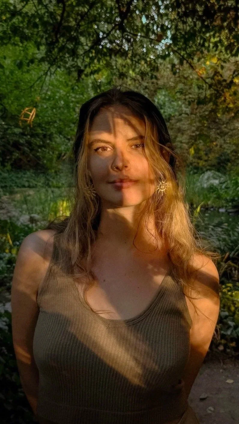 Junge Frau mit langen, gewellten Haaren, trägt ein ärmelloses Oberteil, steht im Freien mit grüner, bewaldeter Umgebung, Sonnenlicht fällt auf ihr Gesicht.