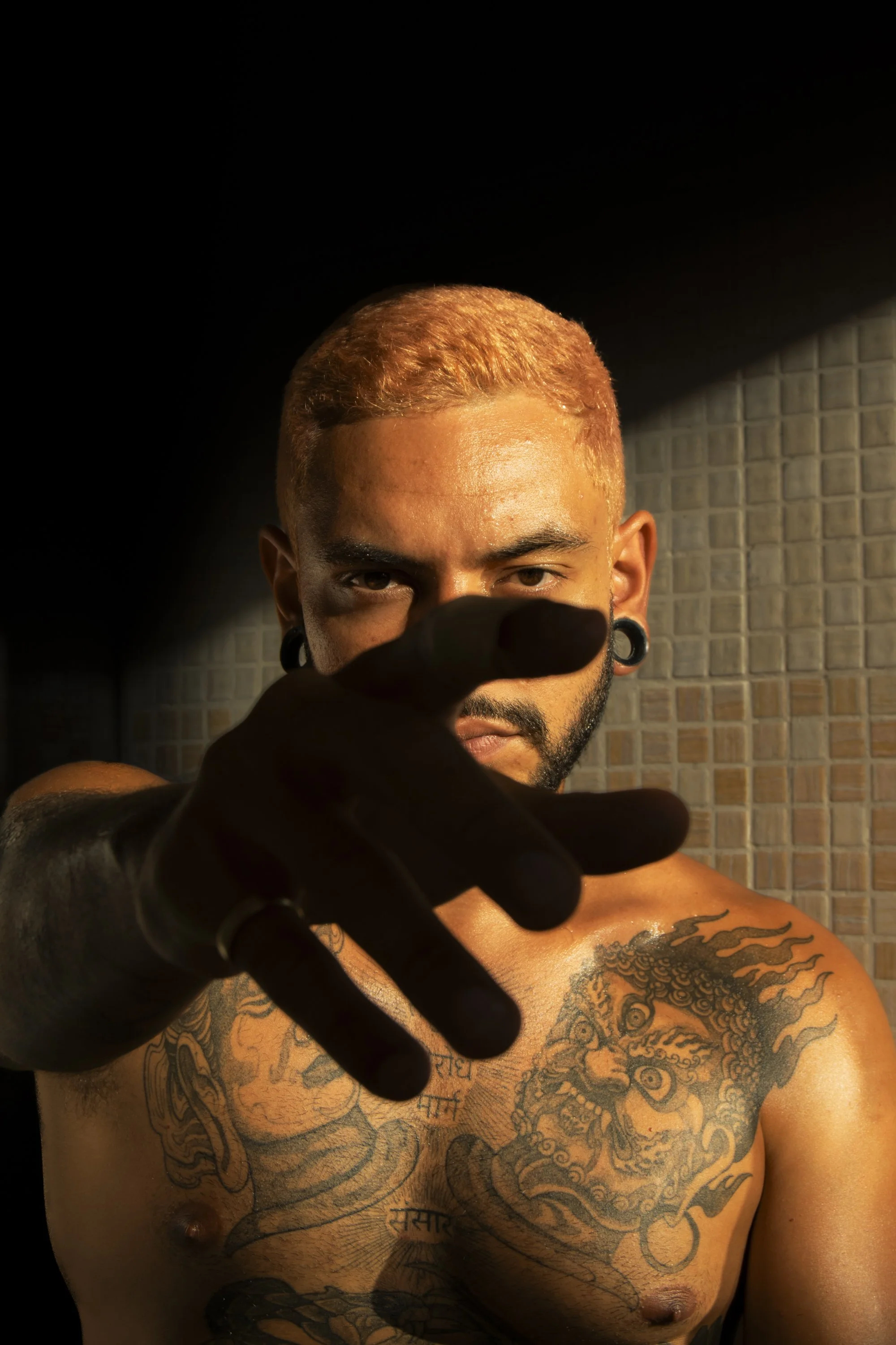 Nahaufnahme eines Mannes mit Tattoos auf der Brust, der in die Kamera blickt und die Hand in die Kamera hält, vor einem dunklen Hintergrund.