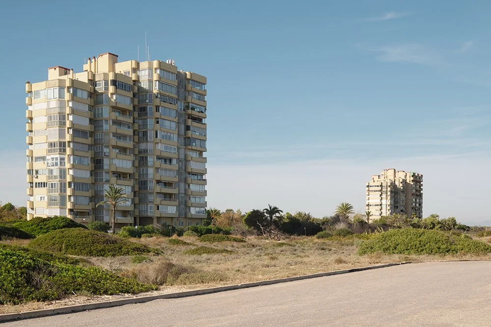 Two tall apartment buildings in a desert-like landscape with sparse greenery and a paved road in the foreground.