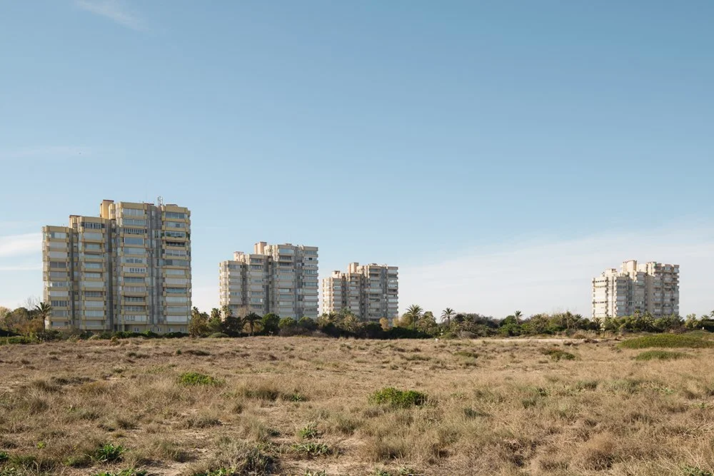 Four tall apartment buildings in the distance with a dry grassy field in the foreground under a blue sky.