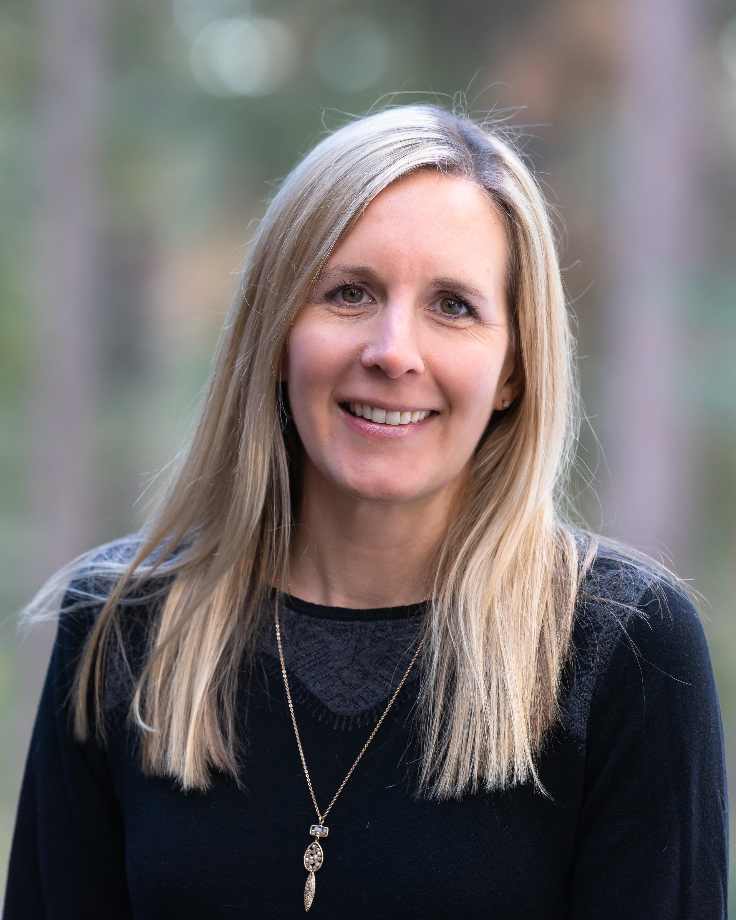 Portrait of a smiling woman with blond hair, wearing a black top and a gold necklace, outdoors with blurred background of trees.