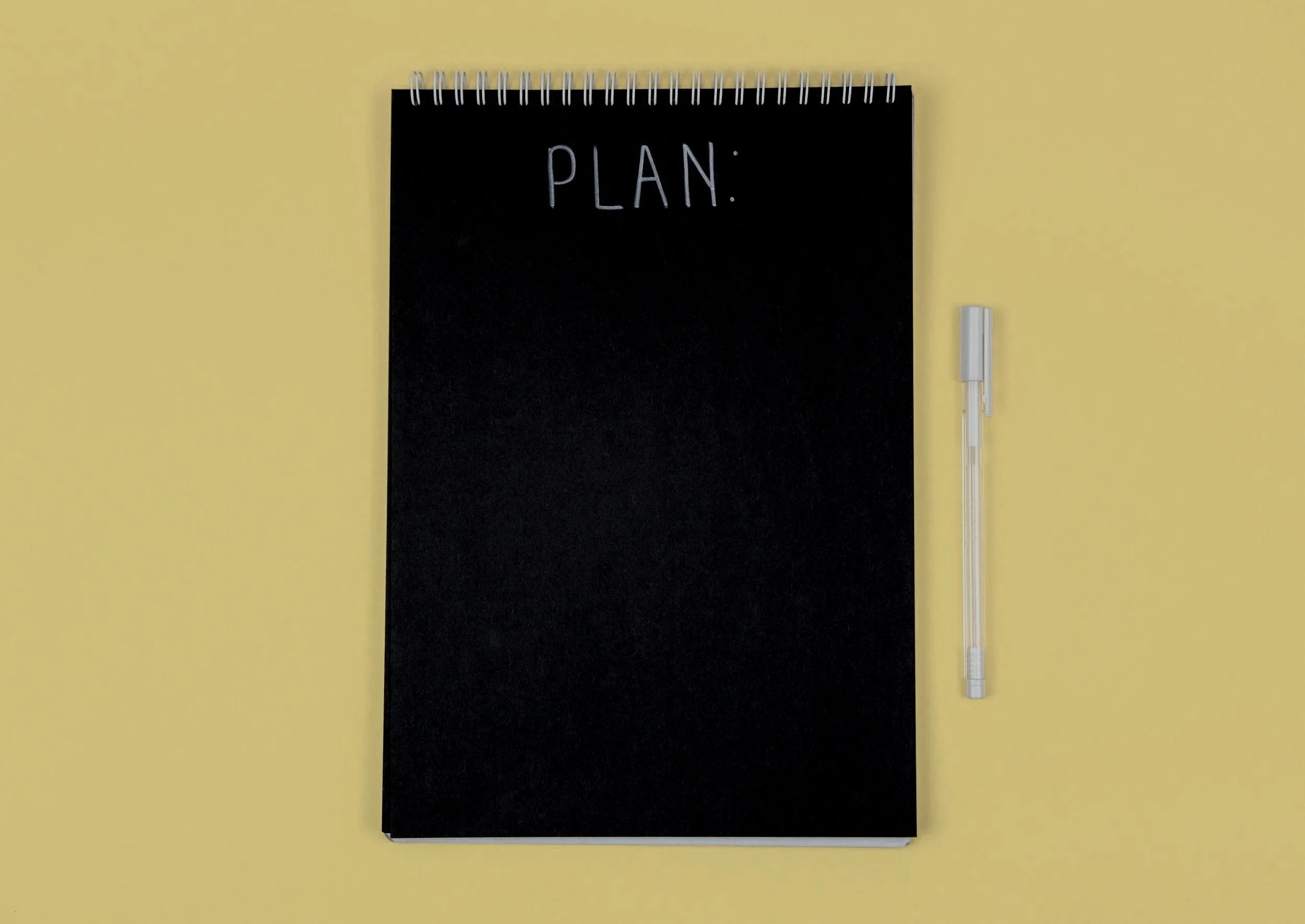 A black spiral-bound notebook with the word 'PLAN:' written on the cover, placed on a yellow surface, with a clear pen beside it.