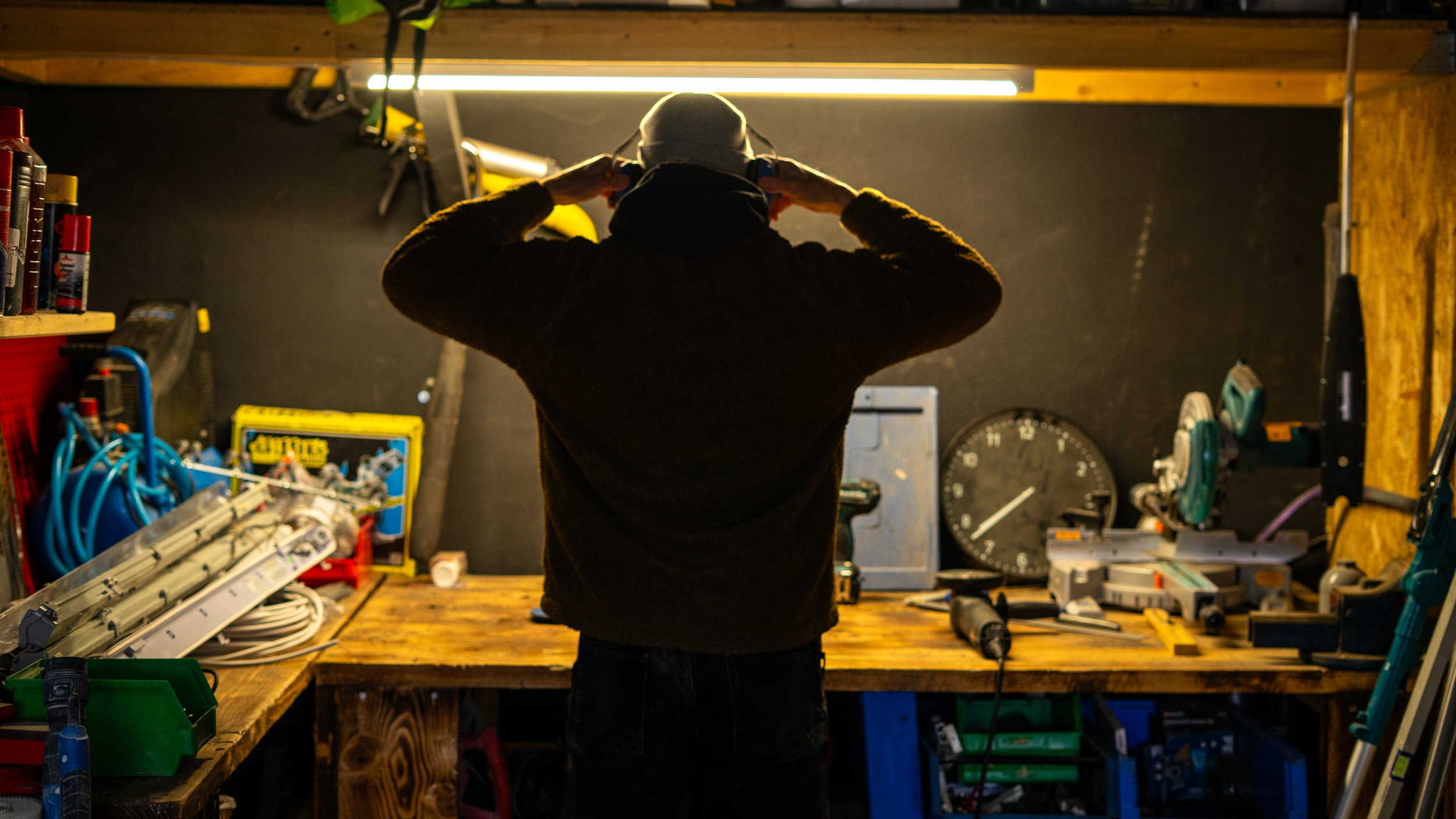 Eine Person steht in einer Werkstatt vor einem Arbeitstisch, die Kamera zeigt die Rückseite der Person, die einen schwarzen Pullover und eine graue Mütze trägt, während sie ihre Hände an den Kopf legt. Der Hintergrund ist mit Werkzeugen, Messgeräten und anderen Werkzeugen ausgestattet.